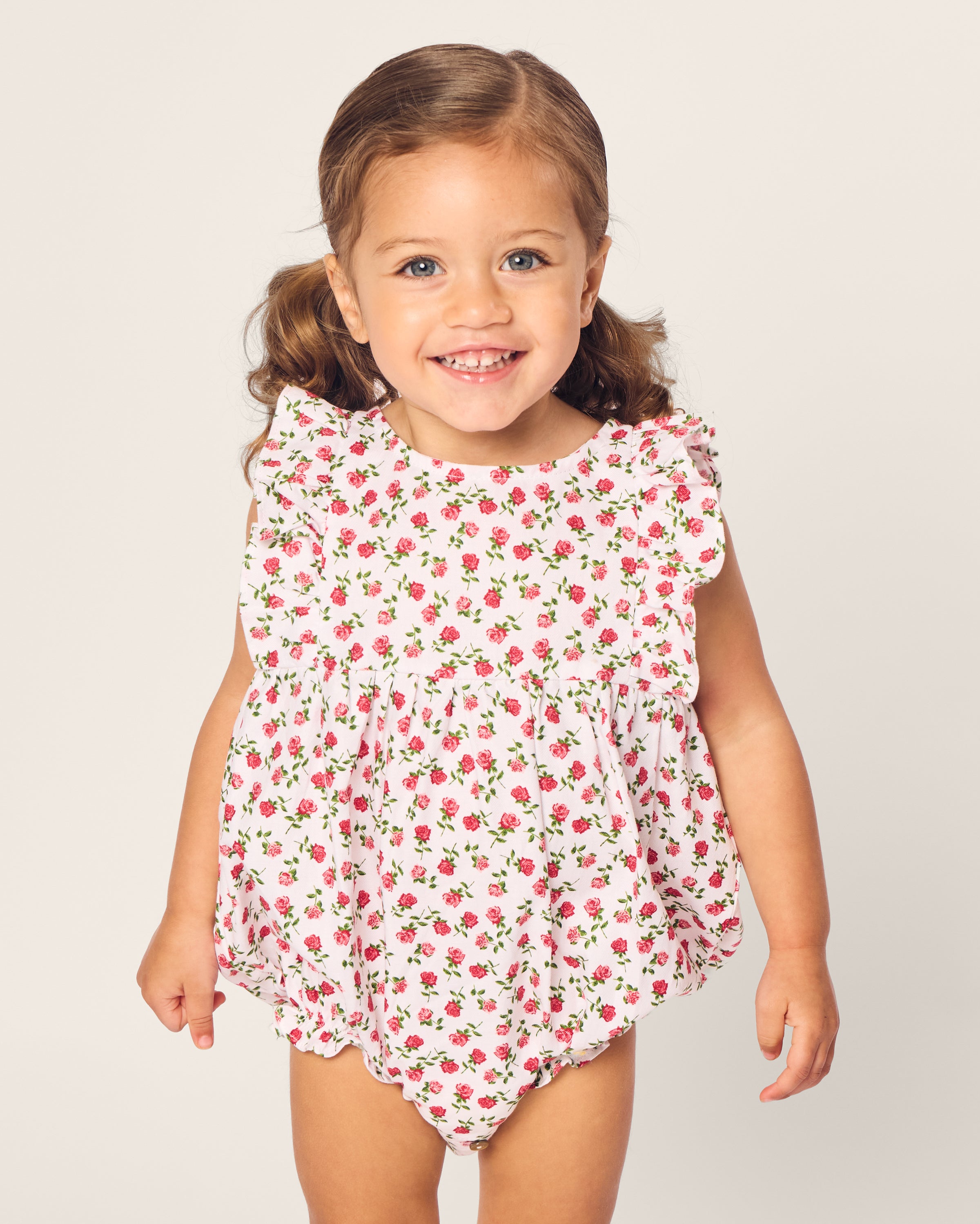 A smiling toddler with brown hair in pigtails wears the Babys Twill Ruffled Romper in Petite Petals by Petite Plume. The luxurious white sleepwear, adorned with pink floral patterns and ruffled sleeves, features yarn-dyed cotton as the child stands against a plain light background.