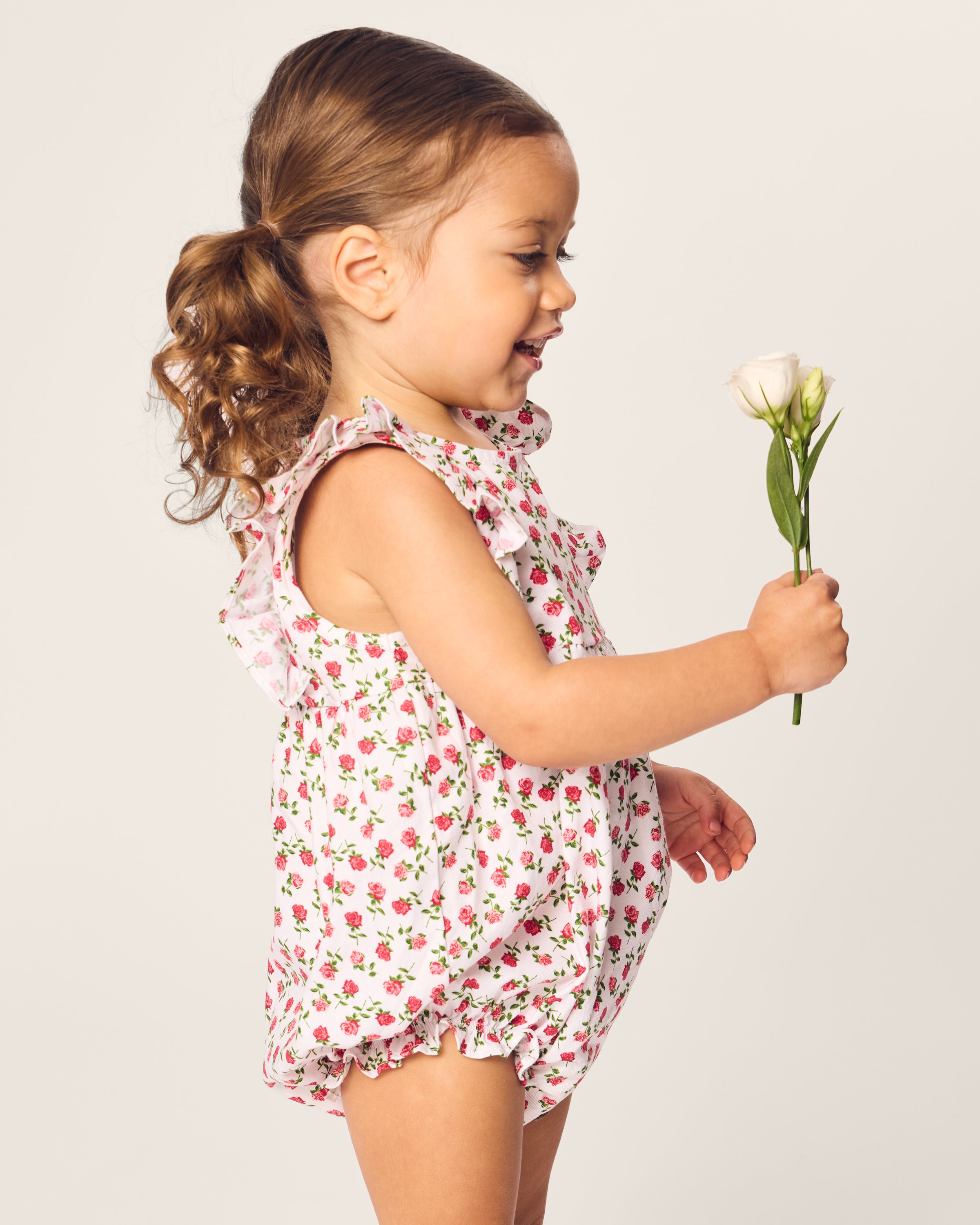 A young girl with curly brown hair, wearing a Petite Plumes Babys Twill Ruffled Romper in Petite Petals, holds a single white rose. She is smiling and facing sideways against a plain background.