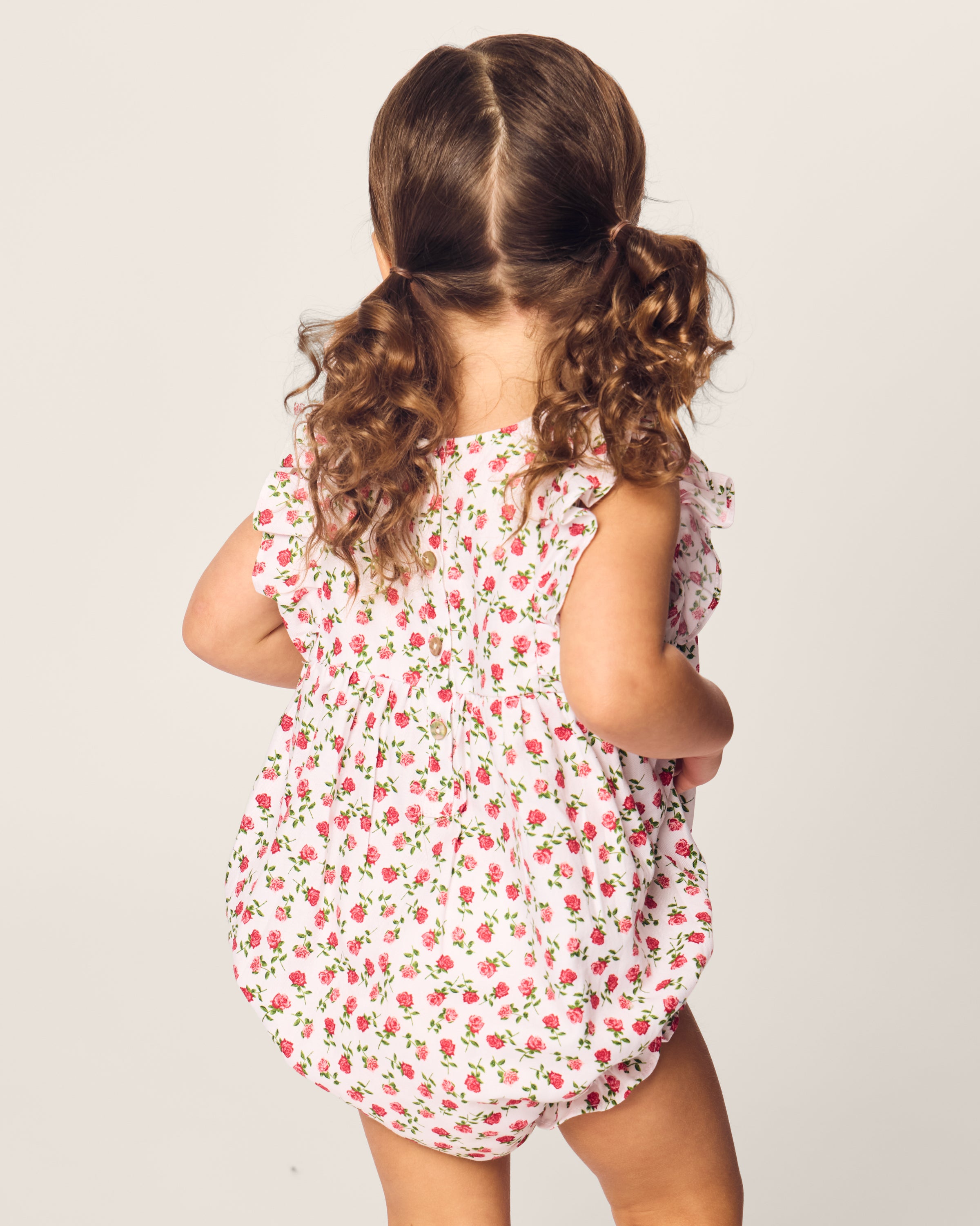 A young child with curly brown hair in pigtails is wearing Petite Plumes Babys Twill Ruffled Romper in Petite Petals. The childs back faces the camera against a plain background.