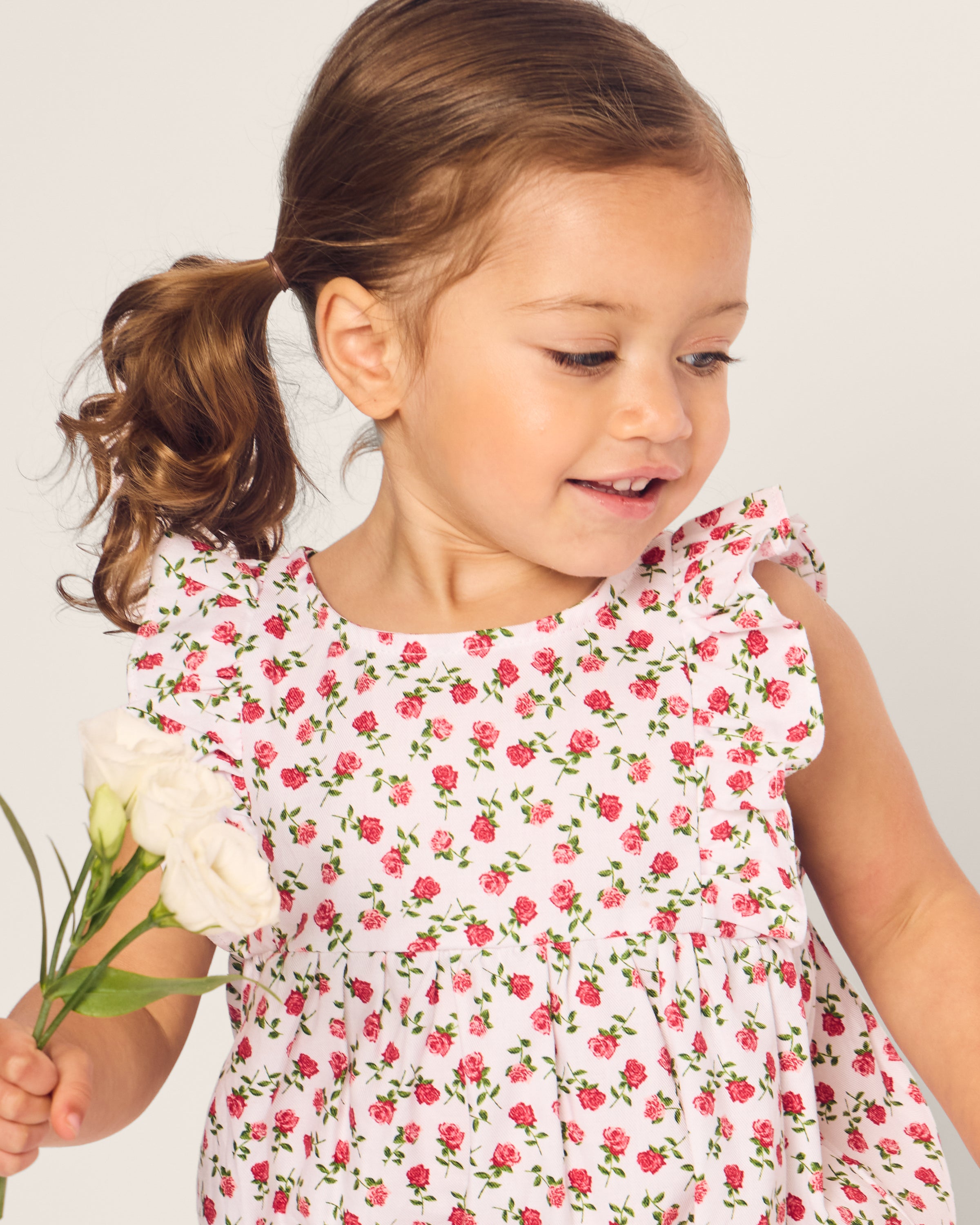 A young girl in Petite Plumes Babys Twill Ruffled Romper in Petite Petals holds a bouquet of white flowers. With her hair in a ponytail, she smiles gently as she looks down. The soft white background enhances the summery, cheerful scene.