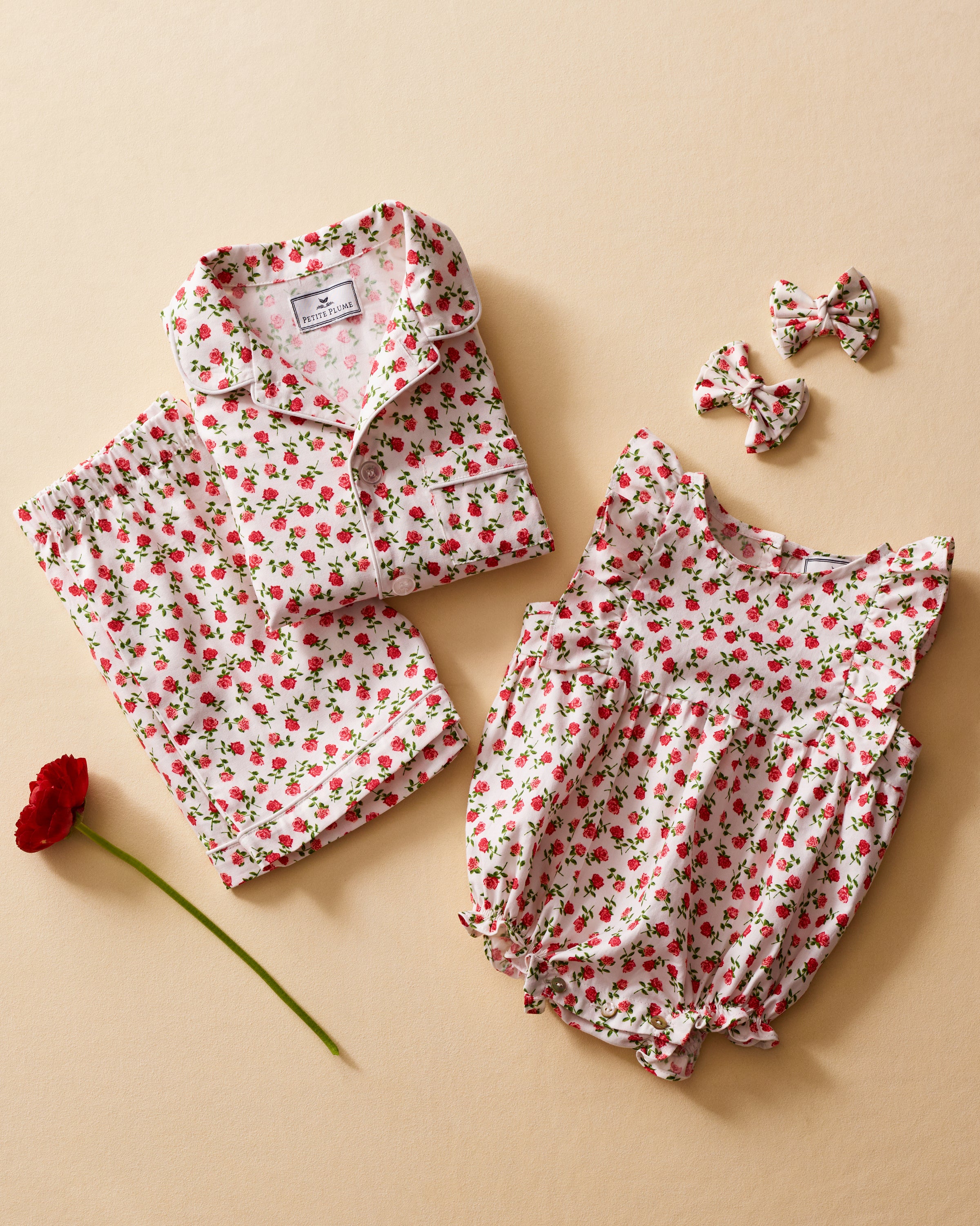 Two floral pajamas from Petite Plume, one is a twill ruffled romper in Petite Petals with pink piping and the other folded on a beige surface, are complemented by matching hair bows and a red rose, inviting restful nights in luxurious sleepwear.