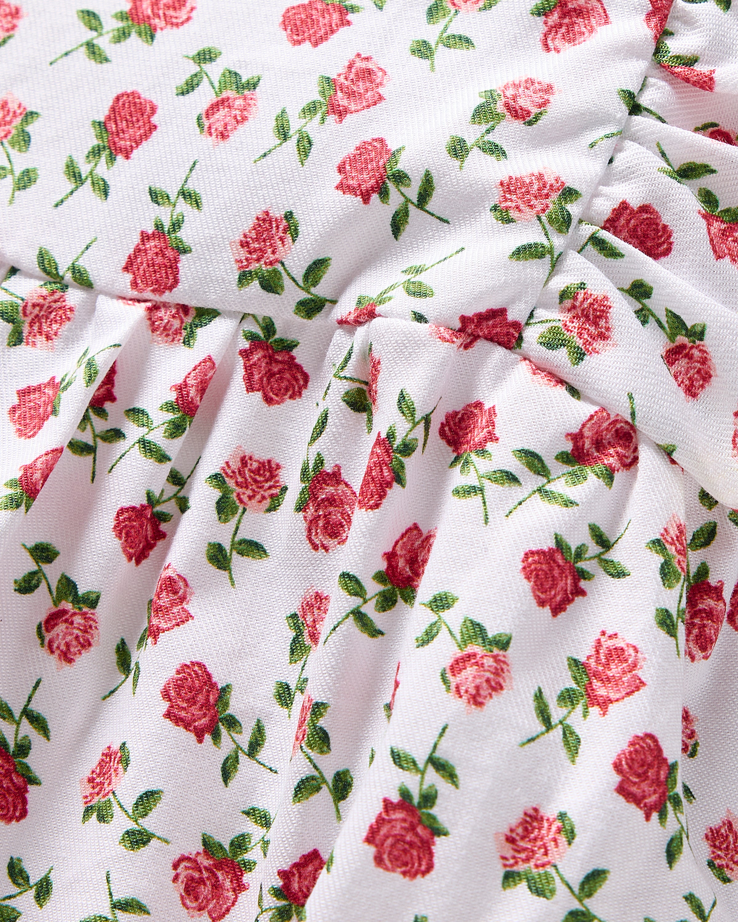 Close-up of Petite Plumes Babys Twill Ruffled Romper featuring a yarn-dyed cotton fabric with petite red roses and green leaves on white. The luxurious material is gathered, offering soft folds and texture perfect for exquisite sleepwear.