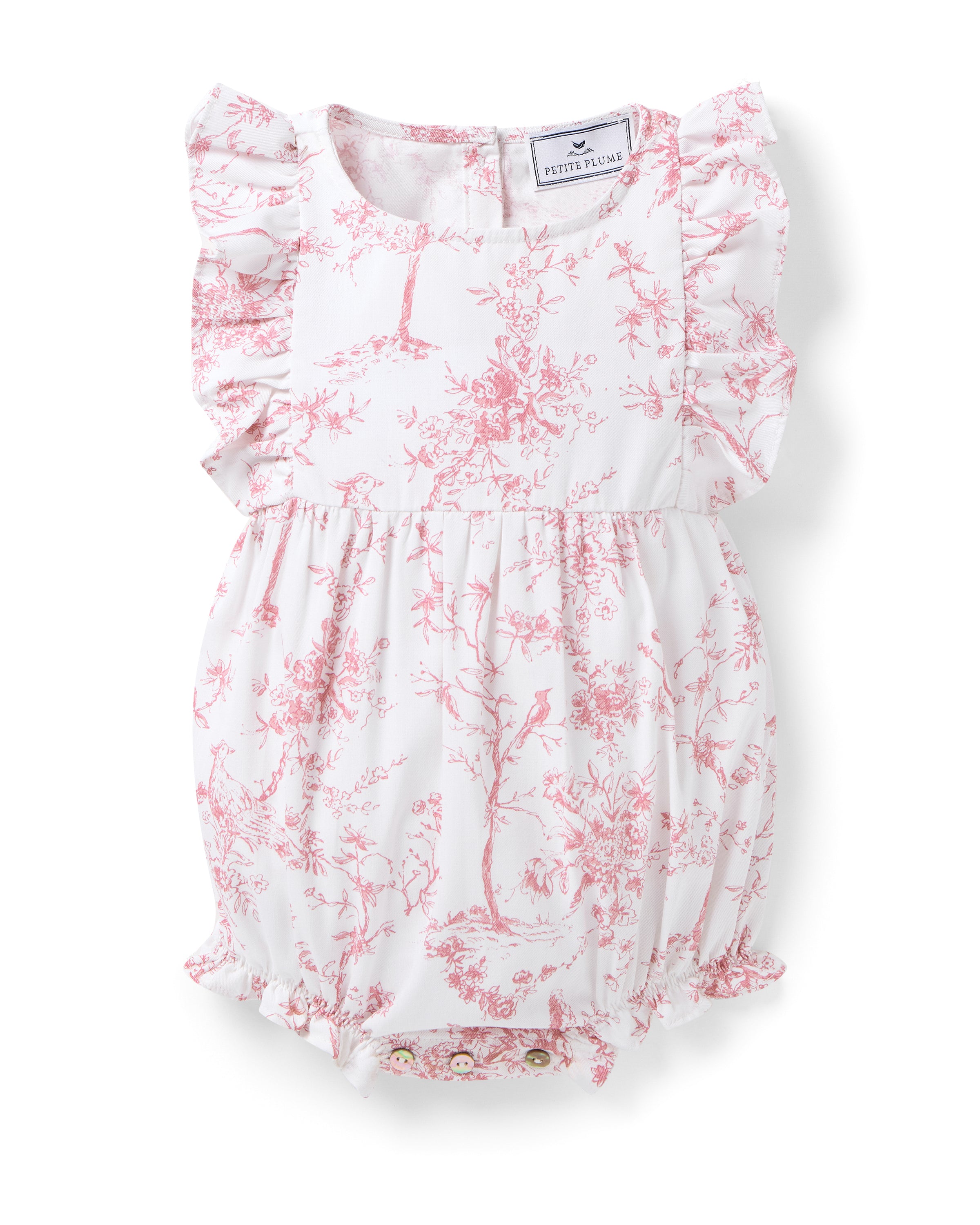 The Pink Timeless Toile Romper by Petite Plume features luxury cotton, ruffled sleeves, and a floral and tree design. Its gathered waist and legs create a bloomer effect for an elegant twist on botanical sleepwear, with a visible tag at the neckline.