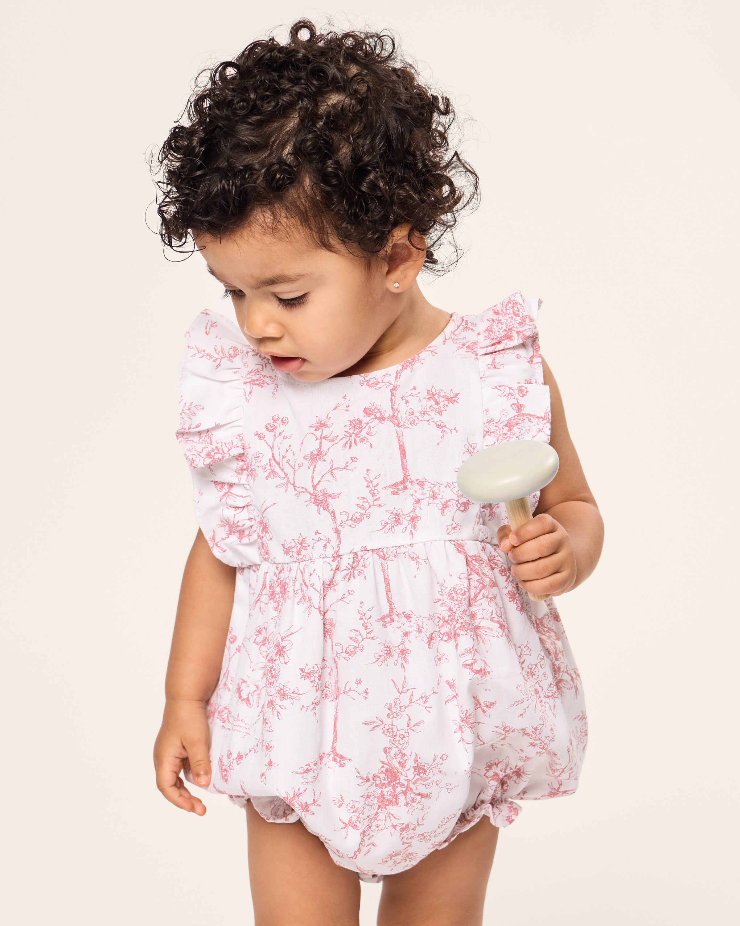 A curly-haired toddler in Petite Plumes Babys Twill Ruffled Romper in Pink Timeless Toile stands against a plain backdrop, holding a small round object. The white luxury cotton fabric features delicate pink floral patterns, adding elegance to the charming scene.