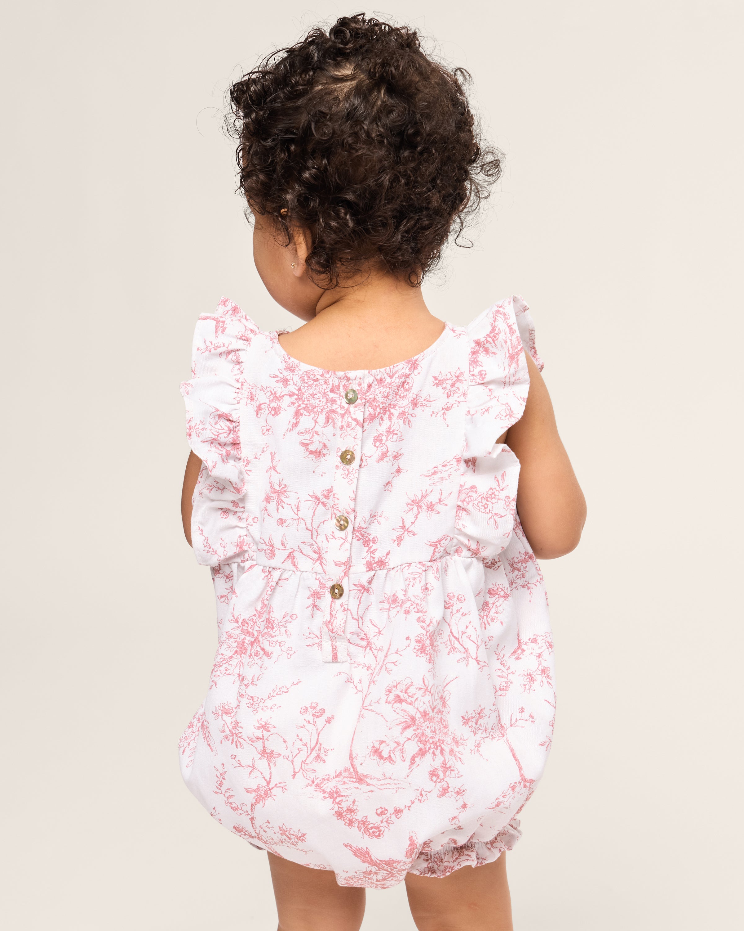 A toddler with curly hair wears Petite Plumes Babys Twill Ruffled Romper in Pink Timeless Toile, featuring floral patterns and ruffled sleeves. The romper has buttons down the back, and the child stands facing away against a plain background.