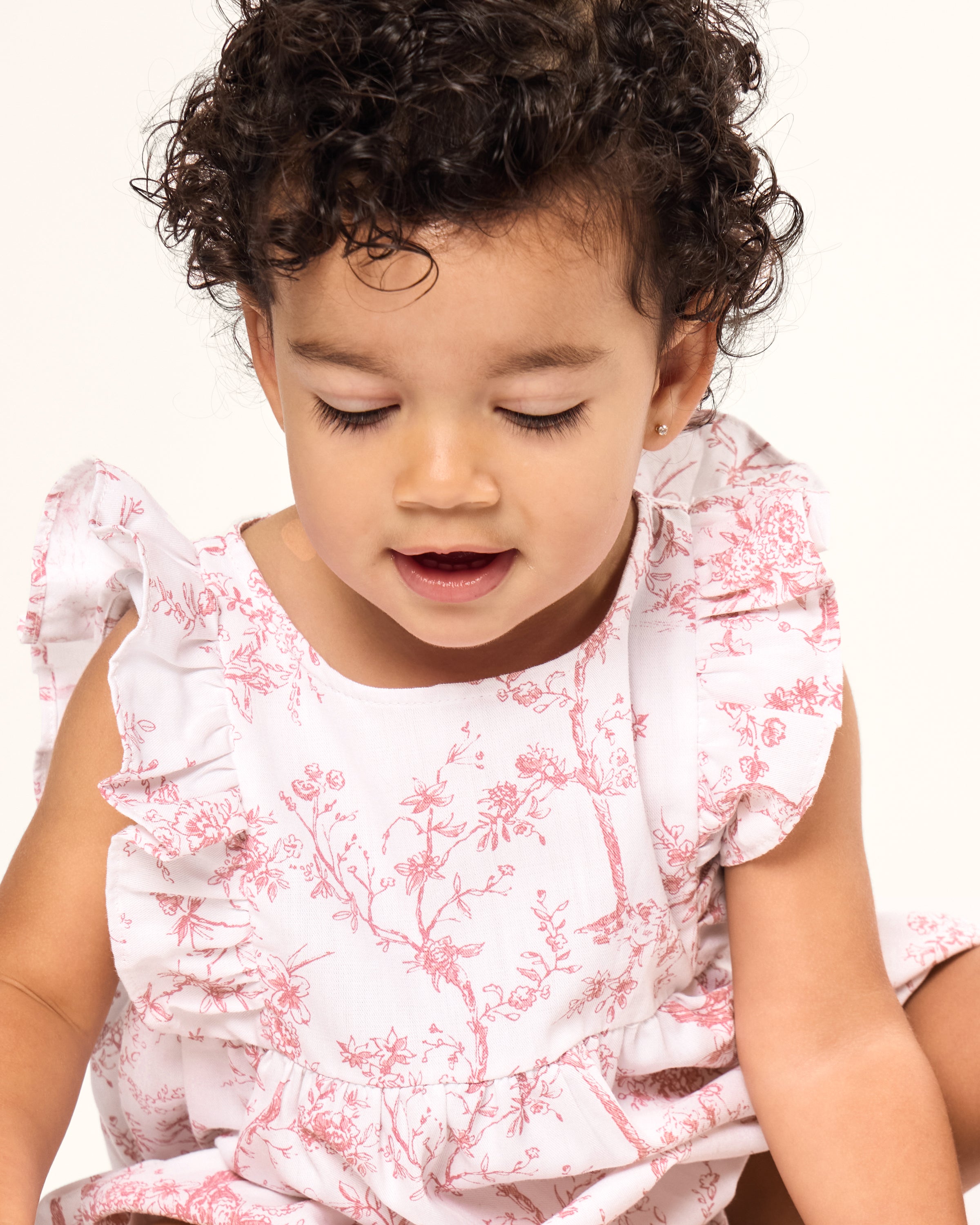 A curly-haired toddler sits serenely, wearing a luxurious Babys Twill Ruffled Romper in Pink Timeless Toile by Petite Plume. Eyes closed with a gentle smile, the childs joyful expression stands out against the softly lit, plain background.