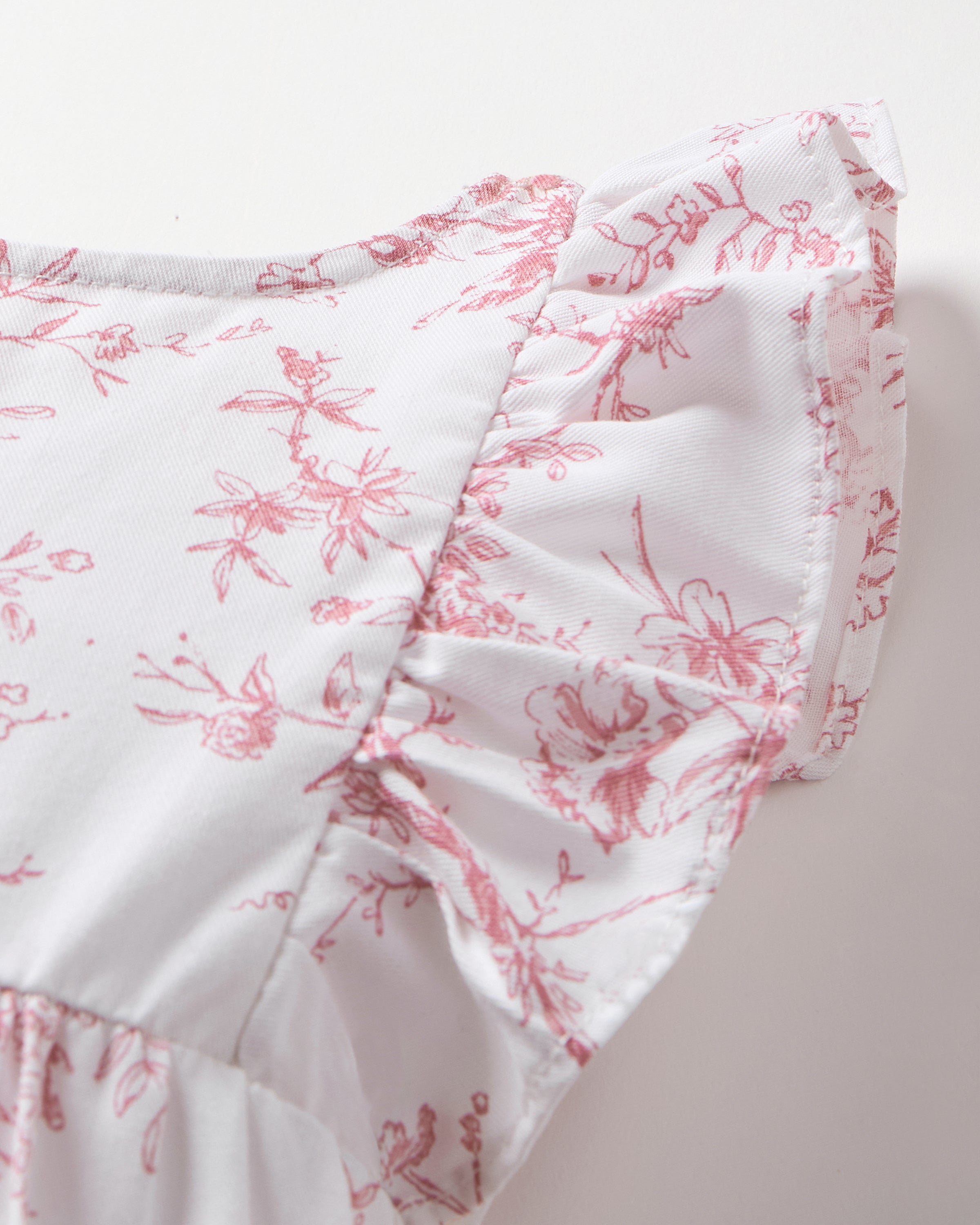 The close-up of Petite Plumes Babys Twill Ruffled Romper in Pink Timeless Toile showcases a white garment with pink floral patterns and delicate edge ruffles. Made from high-quality cotton, its small flowers and leaves create a soft, feminine elegance.