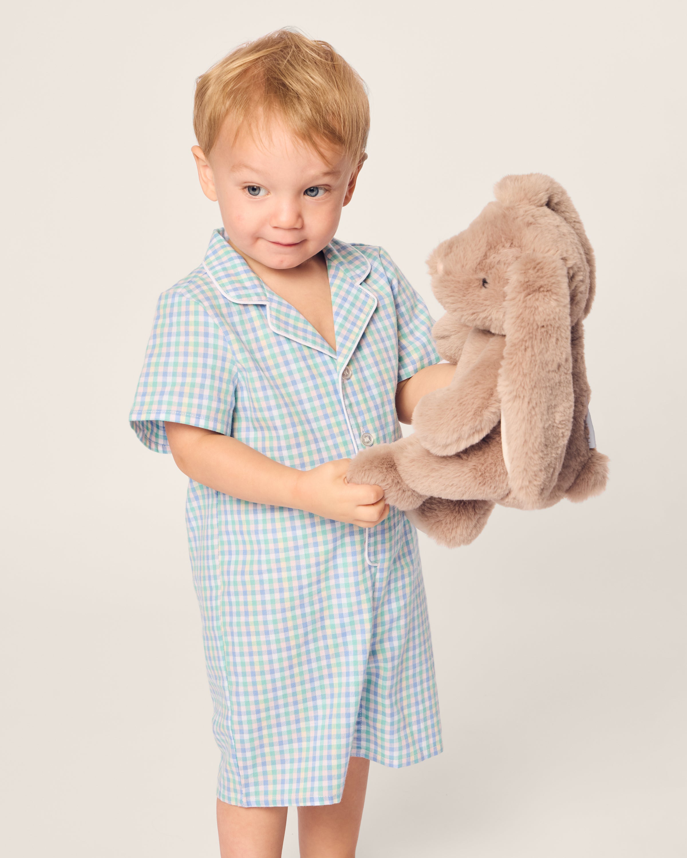 A young child with light brown hair, dressed in Petite Plumes Babys Twill Summer Romper in Spring Gingham, holds a soft brown stuffed bunny against a plain, light-colored background.