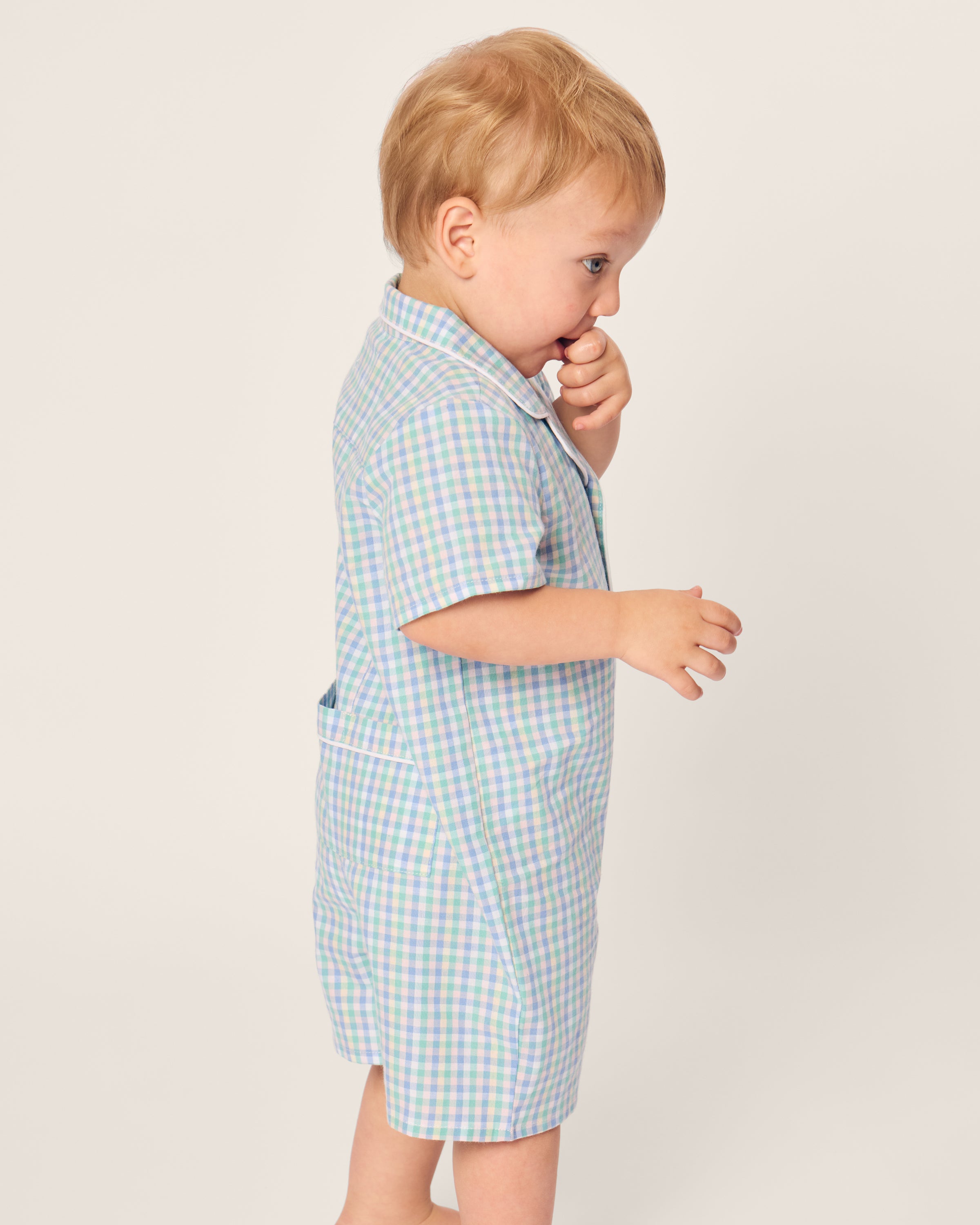 A toddler with blonde hair, wearing Petite Plumes Babys Twill Summer Romper in Spring Gingham, stands sideways. The child touches their mouth with one hand, gazing forward against a plain white background.