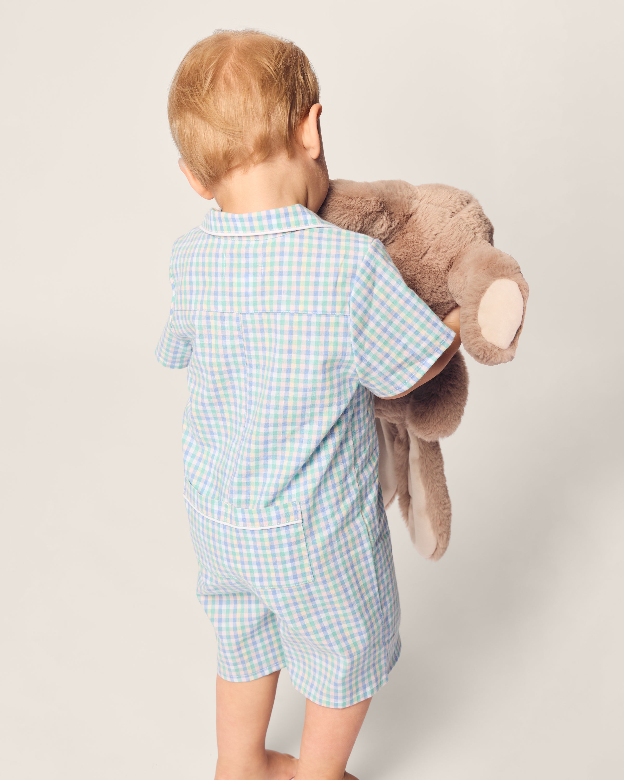 A small child with light hair, wearing Petite Plumes Babys Twill Summer Romper in Spring Gingham, is facing away while holding a large brown stuffed animal against a plain, light-colored background.