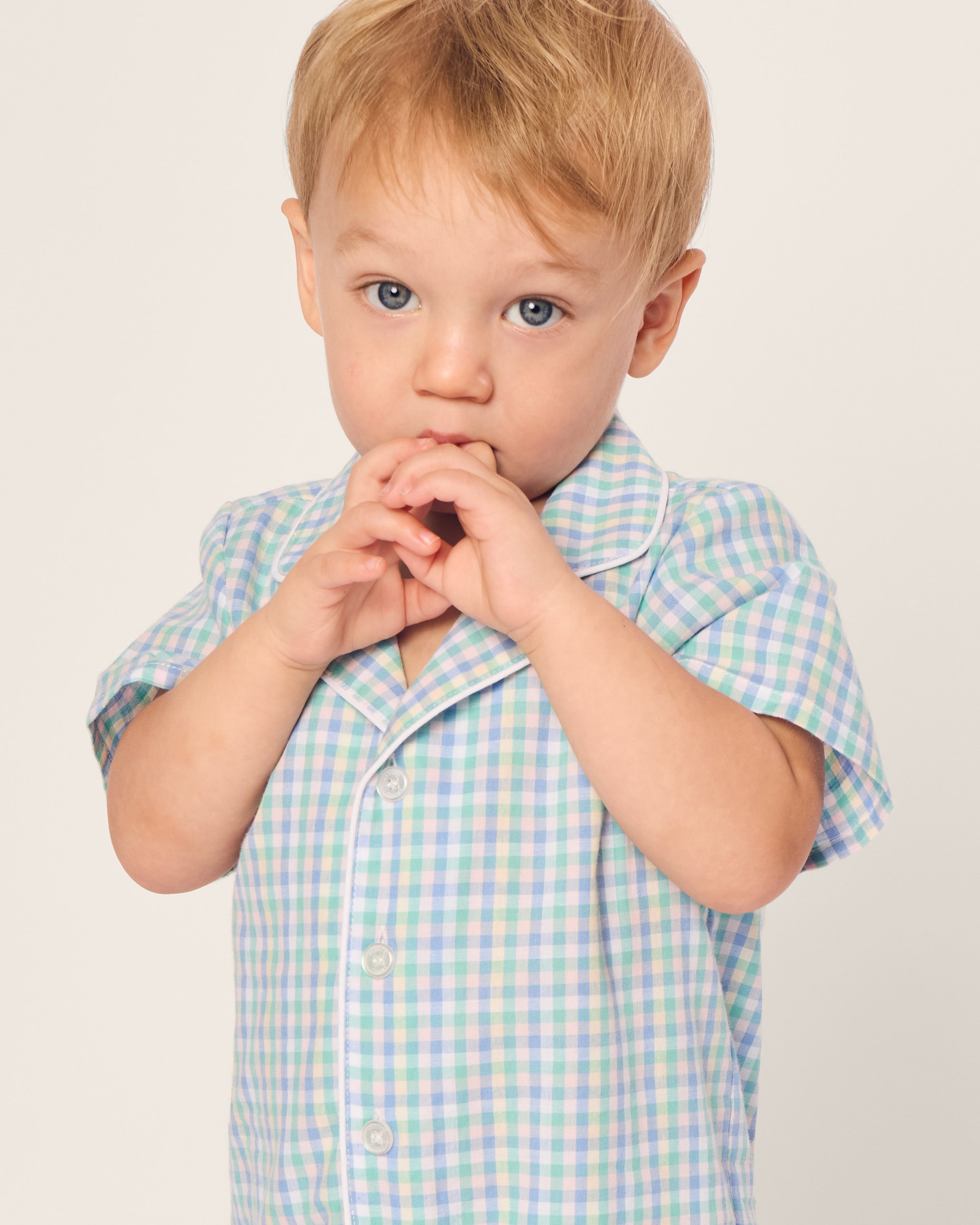 A young child with light hair and a thoughtful expression is wearing Petite Plumes Babys Twill Summer Romper in Spring Gingham. The child has their hands near their face against a neutral background, showcasing the comfort and style of the brushed twill fabric.