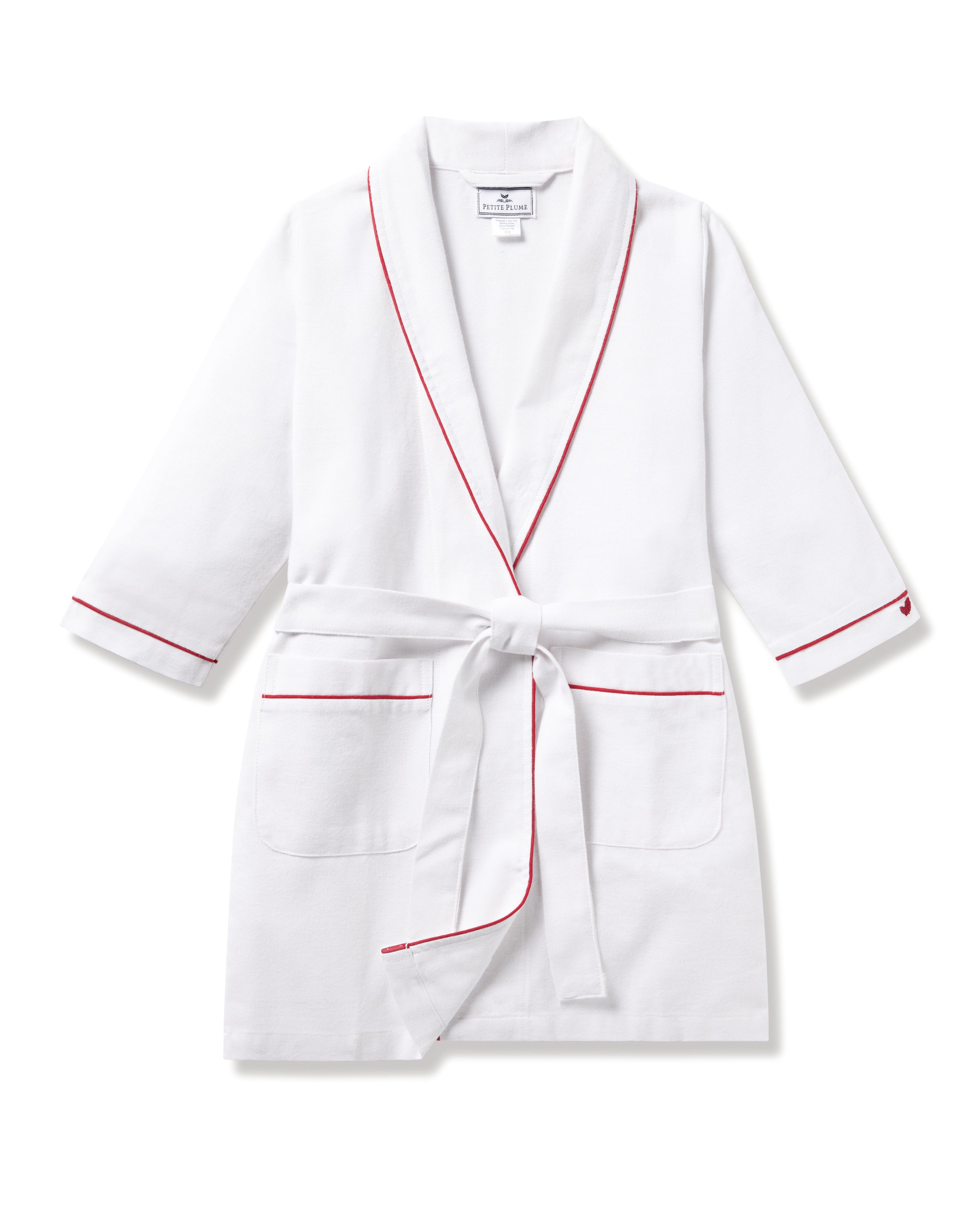 The Kids Flannel Robe in White with Red Piping by Petite Plume is a lightweight childrens sleepwear piece. Featuring a shawl collar, long sleeves, two front pockets, and a matching waist belt, its made from flame-retardant fabric for enhanced safety.