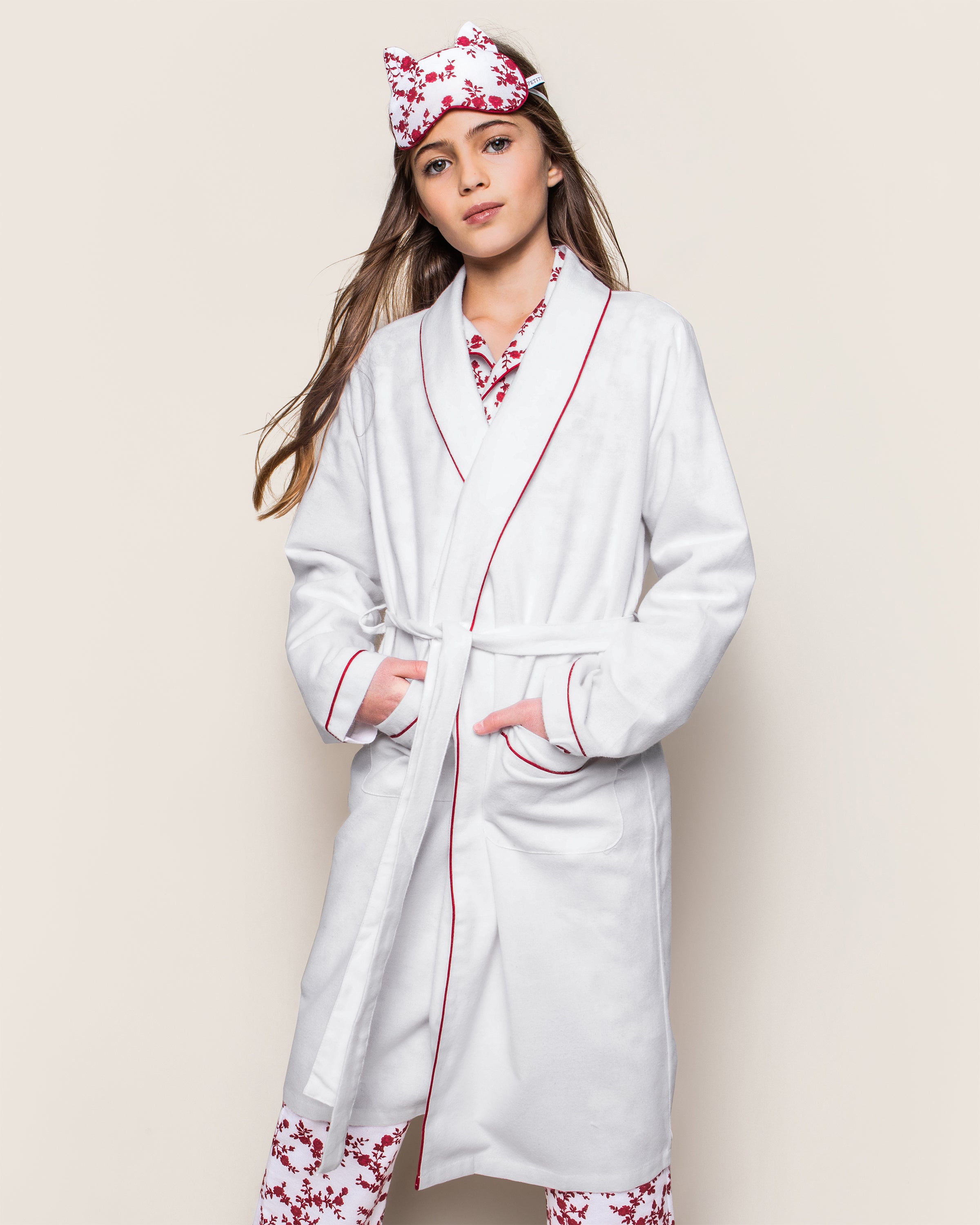 A person poses against a plain background in Petite Plumes Kids Flannel Robe, featuring white fabric with red piping, and wears matching floral sleepwear and headband. The robe has pockets where their hands rest, and their hair flows down.