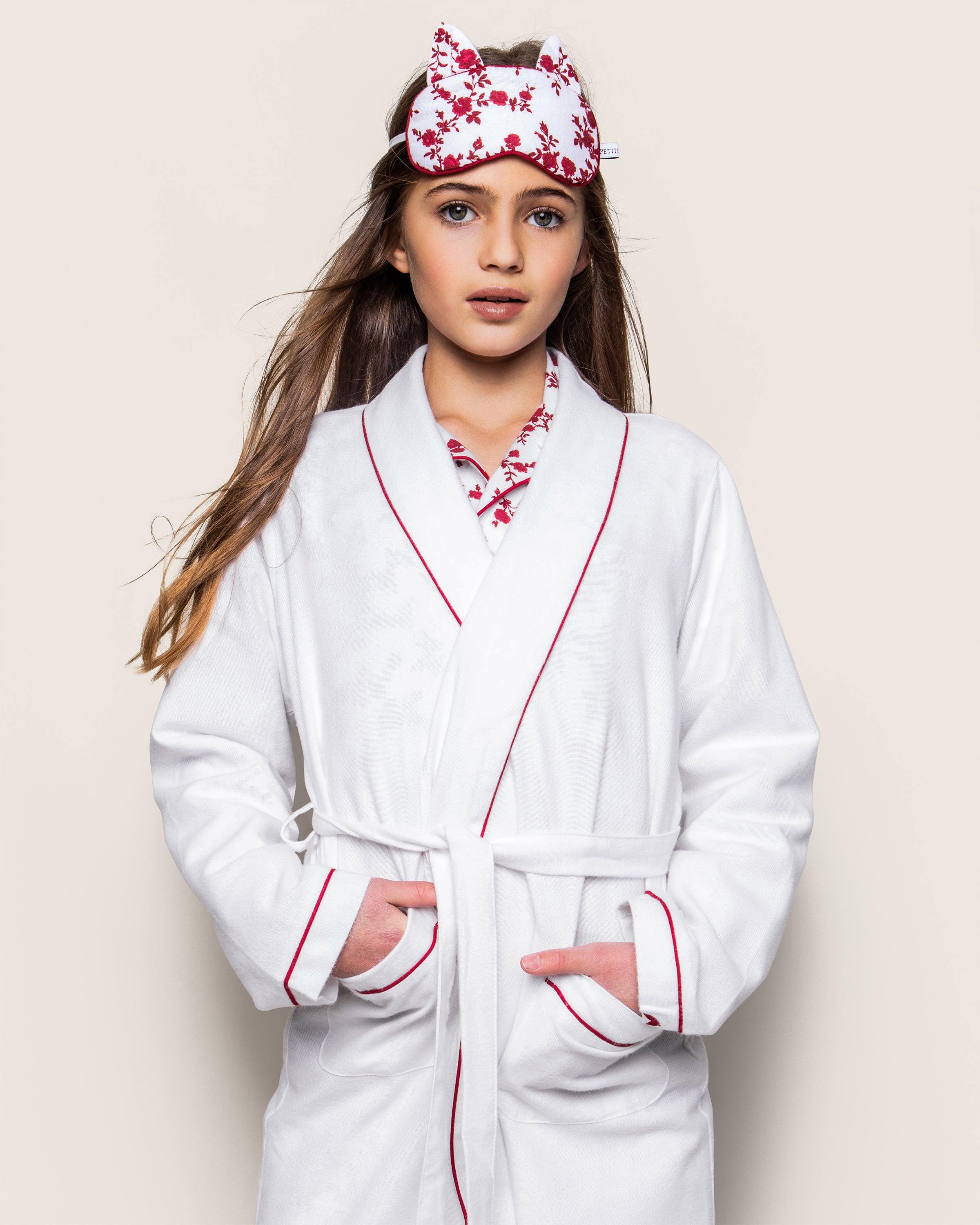 Wearing a Kids Flannel Robe in White with Red Piping by Petite Plume, a long-haired person stands with hands in pockets. Atop their head is a floral sleep mask matching the childrens pajama-inspired top, all set against a plain light backdrop, exuding comfort and ease.