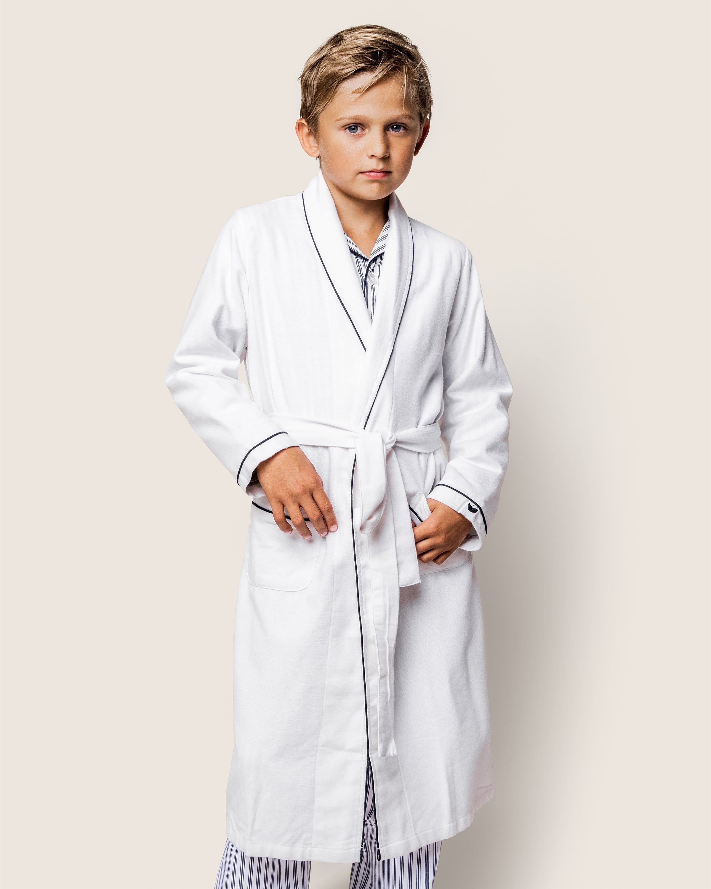 A child with short light brown hair wears a Petite Plume Kids Flannel Robe in White with Navy Piping over striped pajamas, standing against a light background. This flame-retardant robe is tied at the waist as the child gazes neutrally at the camera.