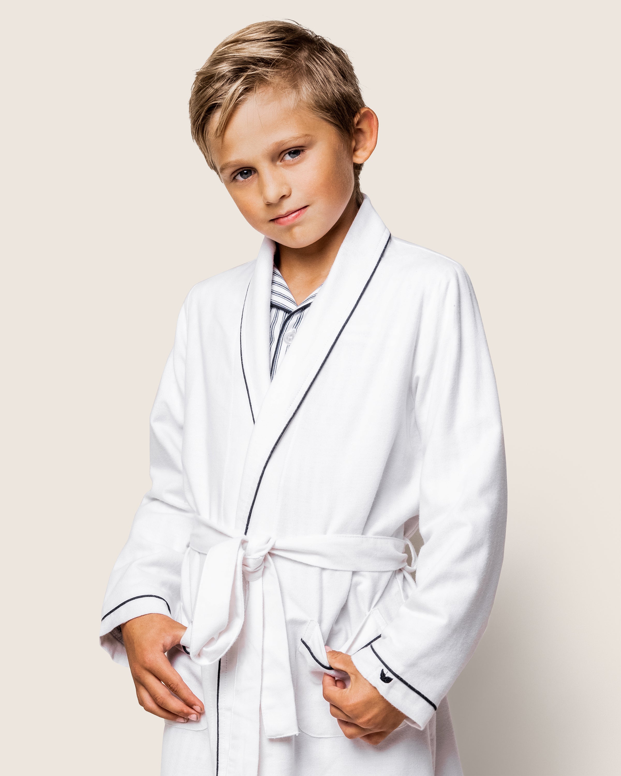 A young blond-haired boy confidently wears the Petite Plume Kids Flannel Robe in White with Navy Piping, gripping its belt and boasting a slight smile against a plain, light-colored background.