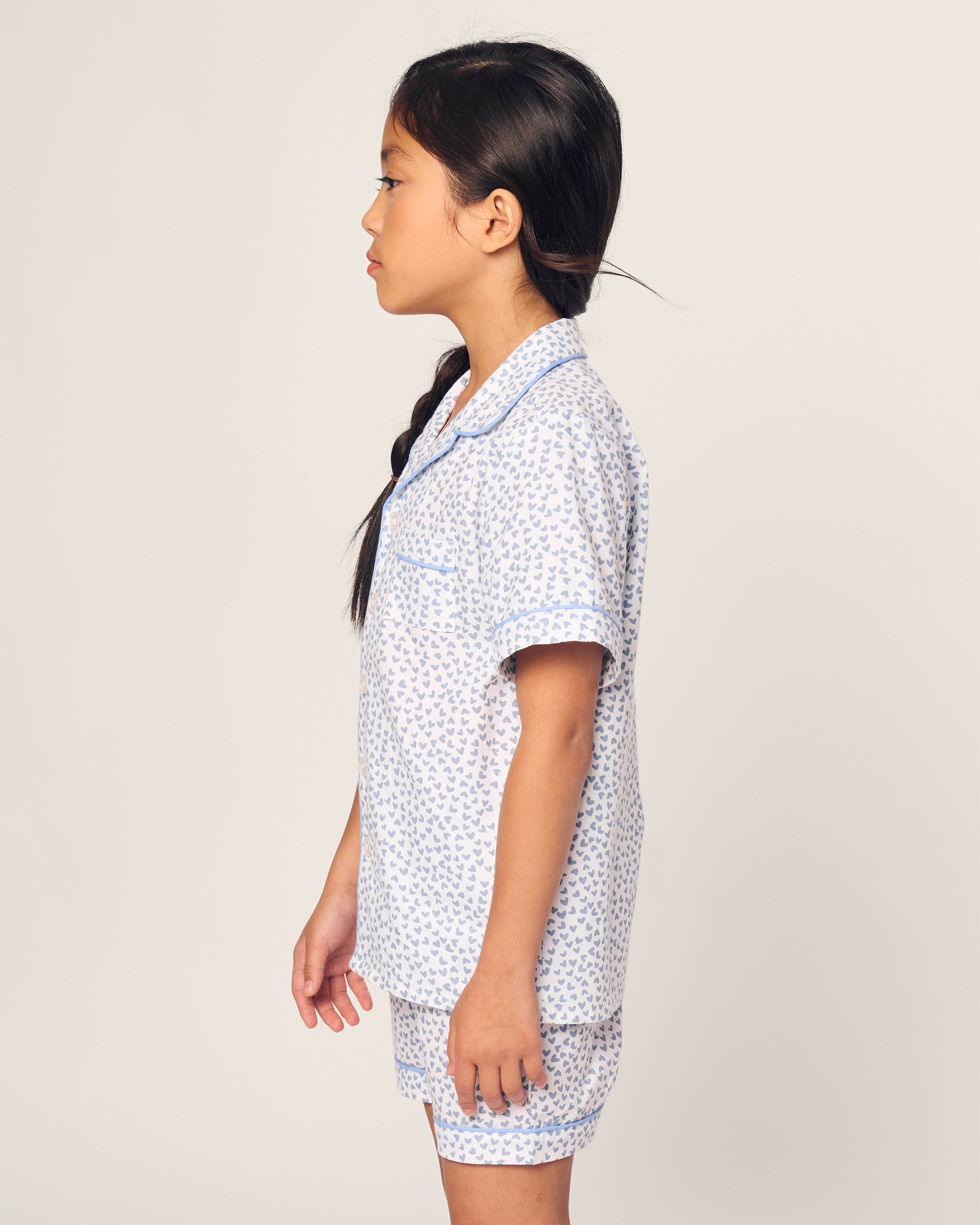 A young girl with long hair tied in a braid stands in profile, wearing Petite Plumes luxurious Kids Twill Short Set in Bluehearts. Her light-colored pajamas feature the delicate design, and the plain background highlights her serene side profile.