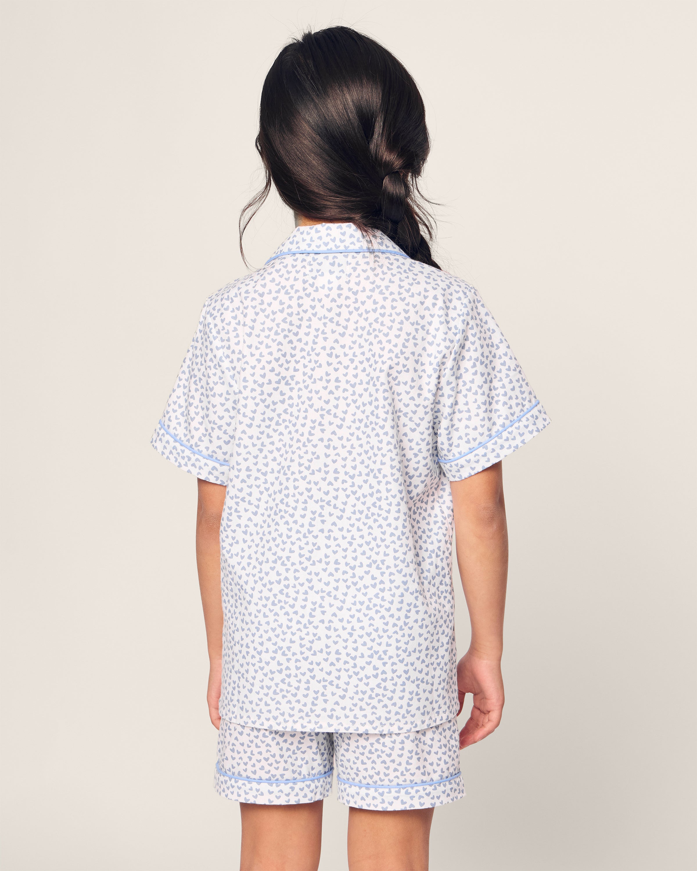 A child with long dark hair styled to one side wears the Petite Plume Kids Twill Short Set in Bluehearts, a luxurious sleepwear set featuring short sleeves and shorts in light blue and white patterns, set against a plain background.