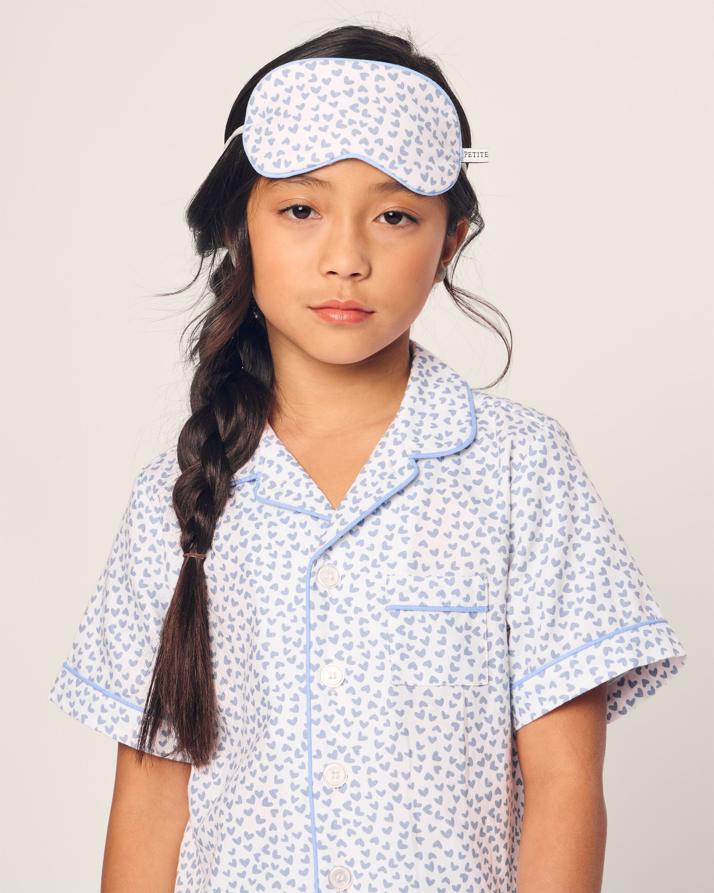 A young girl with a long braid is wearing Petite Plumes Kids Twill Short Set in Bluehearts pajamas. Her matching sleep mask rests above her forehead, highlighting the luxurious sleepwear against a plain background and emphasizing her calm expression.