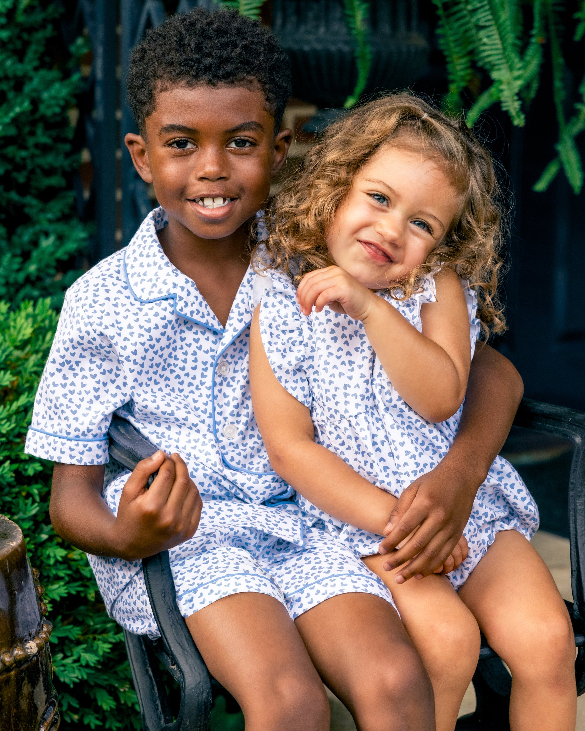 A young boy and girl sit together on a chair, dressed in matching Petite Plumes Kids Twill Short Set in Bluehearts. The boy smiles with his arm around the girl, who shares his happiness. Greenery provides a serene backdrop to their moment of luxury and comfort.