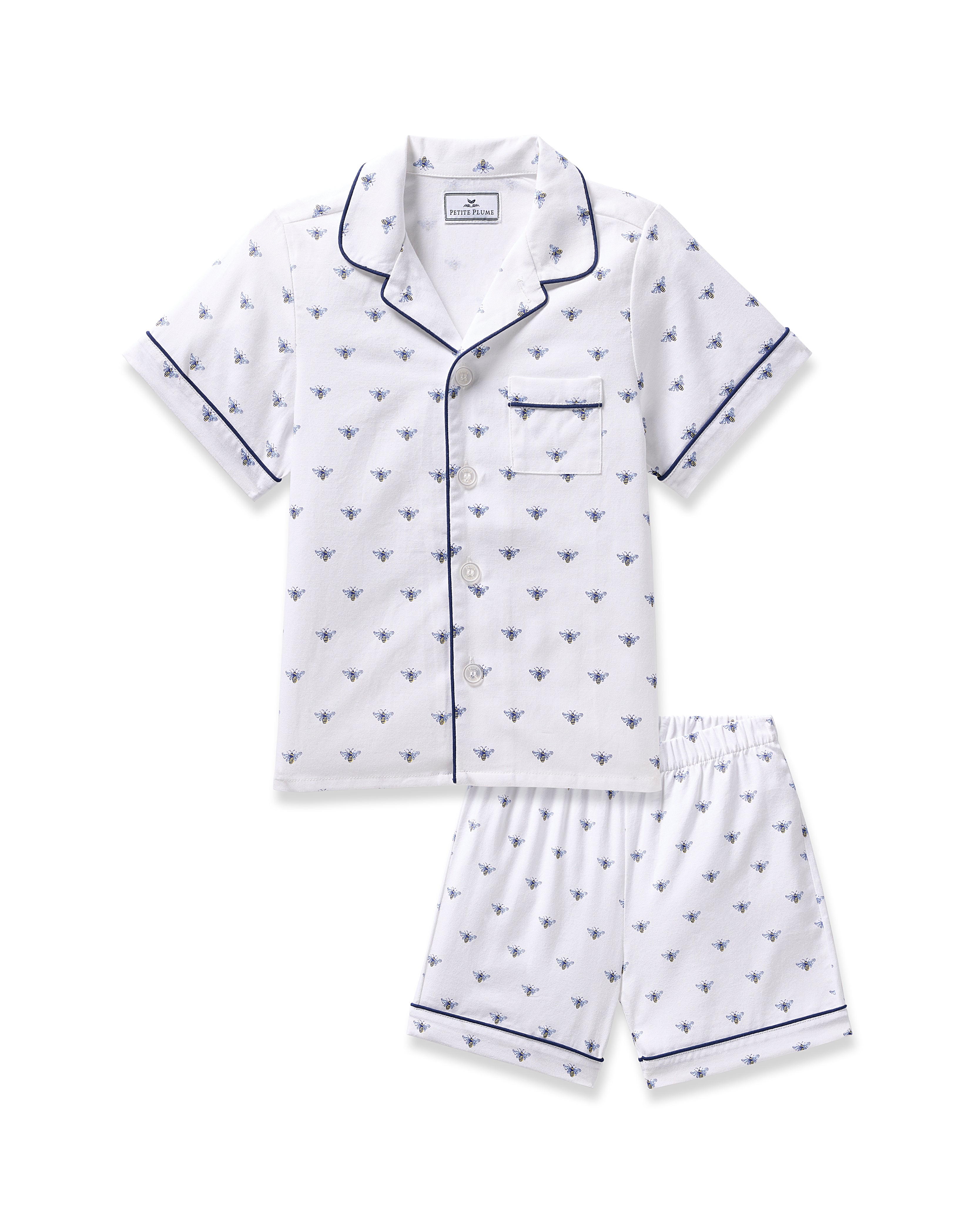 The Kids Twill Short Sleeve Short Set by Petite Plume features a white pajama outfit in premium cotton with a charming Buzzing Bees print, including a short-sleeved button-up shirt, chest pocket, and shorts, all elegantly finished with dark piping.