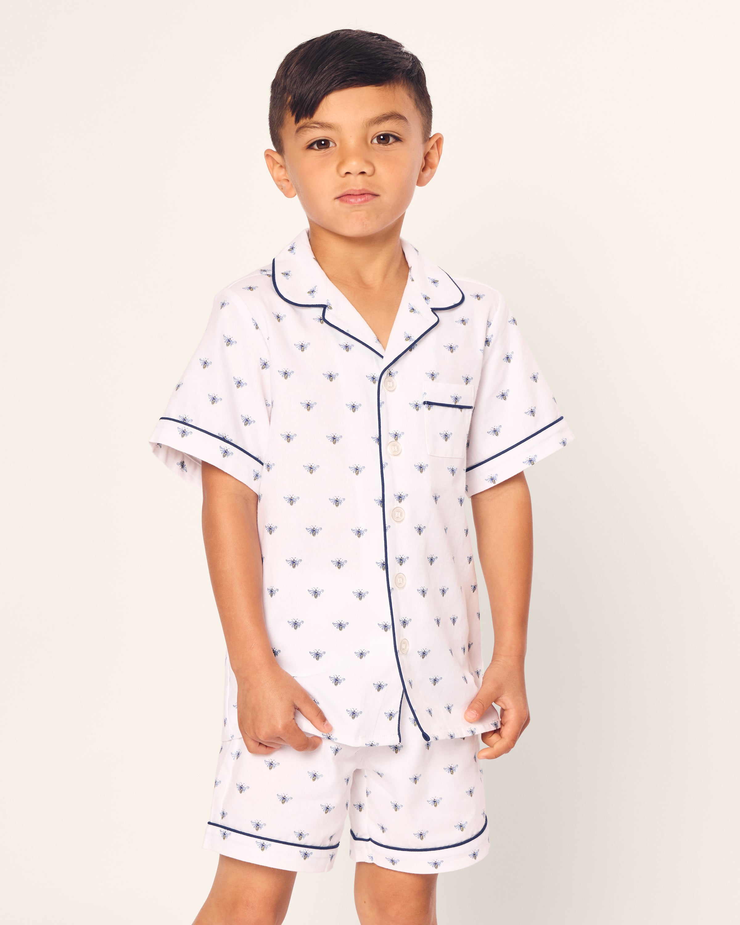 A young boy wears Petite Plumes Kids Twill Short Sleeve Short Set in Buzzing Bees, featuring white pajamas with buzz bee print and navy piping. The set has a button-up top and matching shorts, exuding classic comfort and style against a light background.