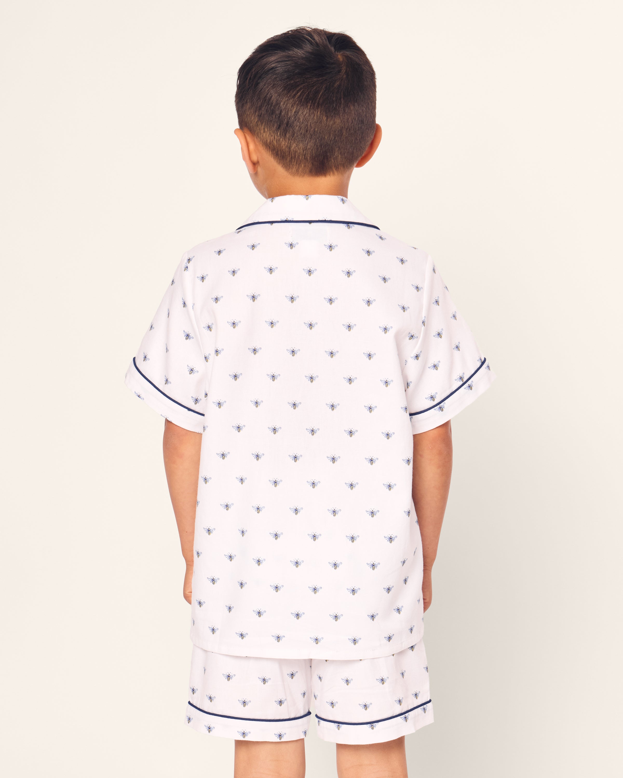 A young boy, seen from the back, wears Petite Plumes luxurious Kids Twill Short Sleeve Short Set in Buzzing Bees print. These white pajamas feature small blue bees and short sleeves accented with blue piping.