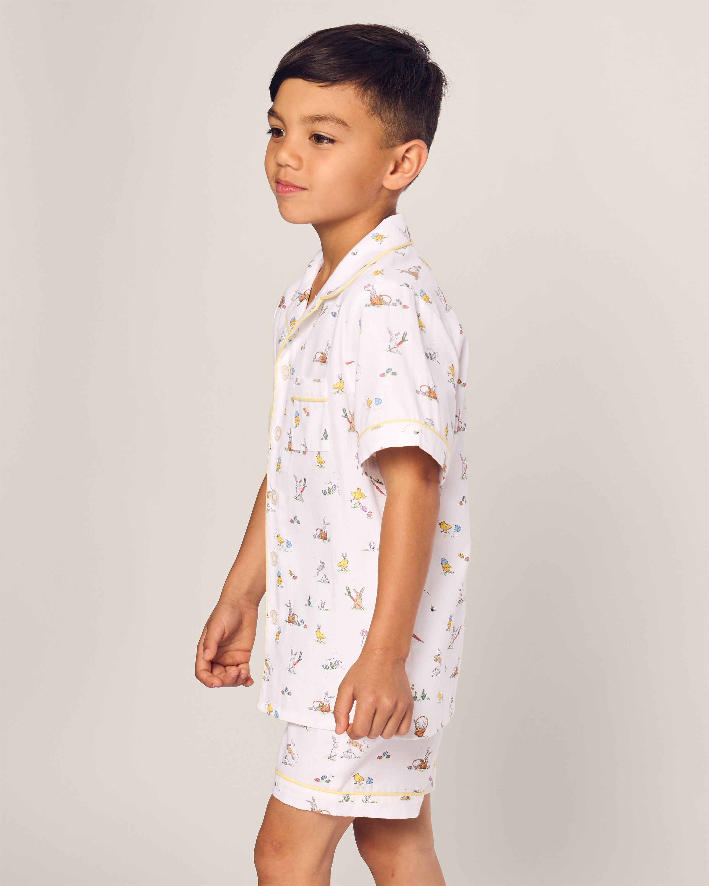 A young boy in side profile faces left, wearing the Petite Plume Kids Twill Short Set. This luxurious sleepwear features white short-sleeved pajamas adorned with a colorful Easter Frolic print on a neutral background.