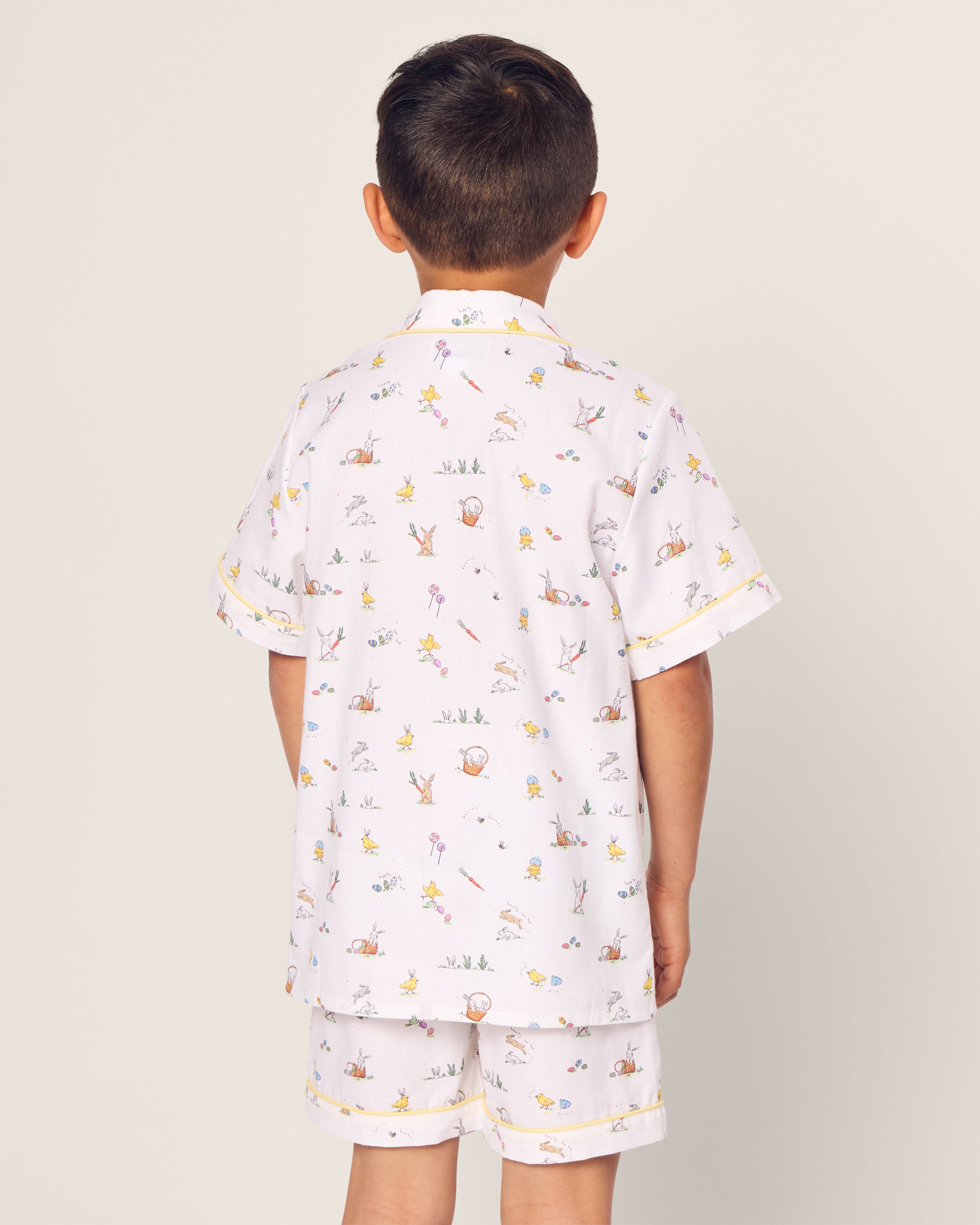 A young boy with short dark hair, his back to the camera, wears Petite Plumes Kids Twill Short Set in Easter Frolic, a luxurious light-colored sleepwear featuring whimsical prints of small animals and sports equipment for perfect comfort.