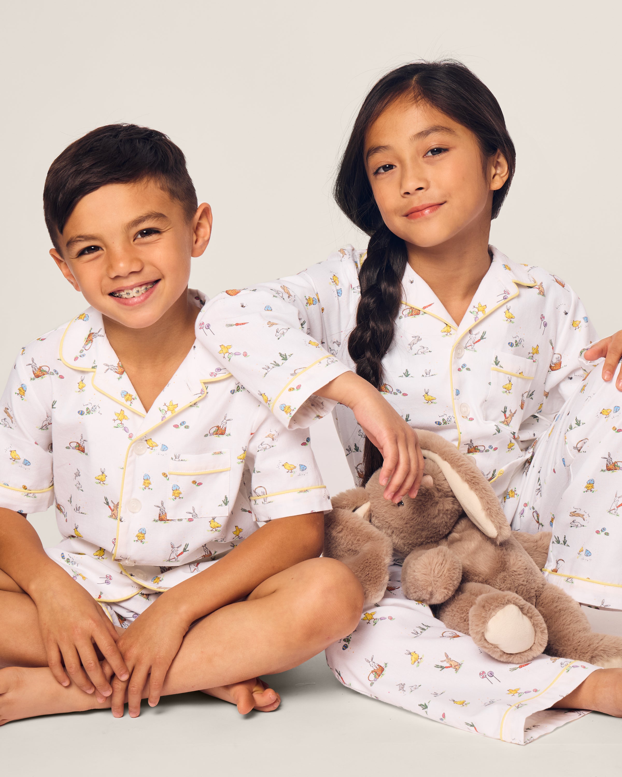 Two smiling children in luxurious Petite Plume sleepwear sit on the floor. They sport matching Kids Twill Short Sets in an Easter Frolic print. One child cuddles a stuffed bunny, embodying comfort and joy against a plain backdrop.