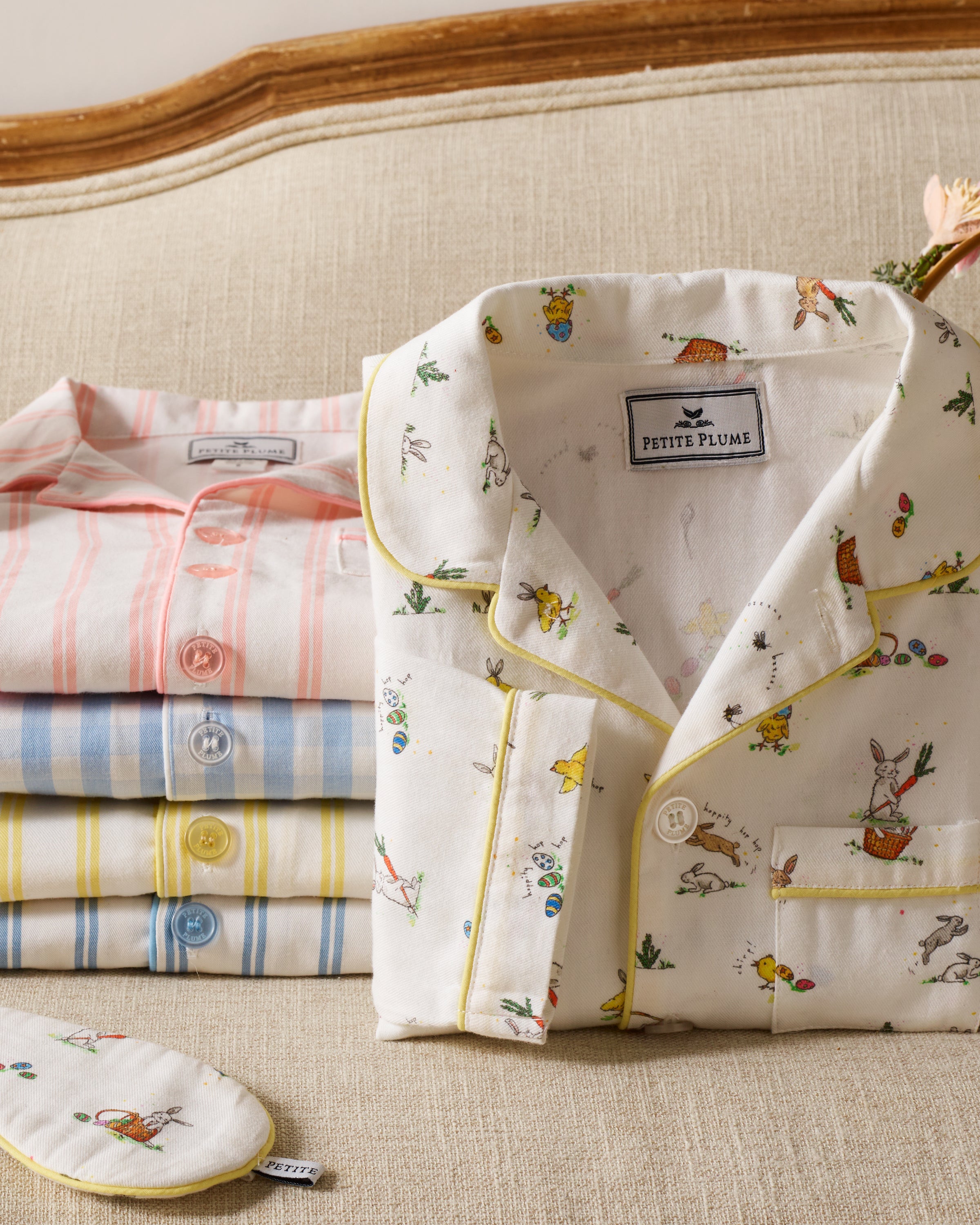 Four neatly folded pairs of luxurious sleepwear rest on a beige sofa. The top pair is the Kids Twill Short Set in Easter Frolic from Petite Plume, featuring yellow piping. Others include striped designs in pink, blue, and yellow. An eye mask with matching print sits to the left.