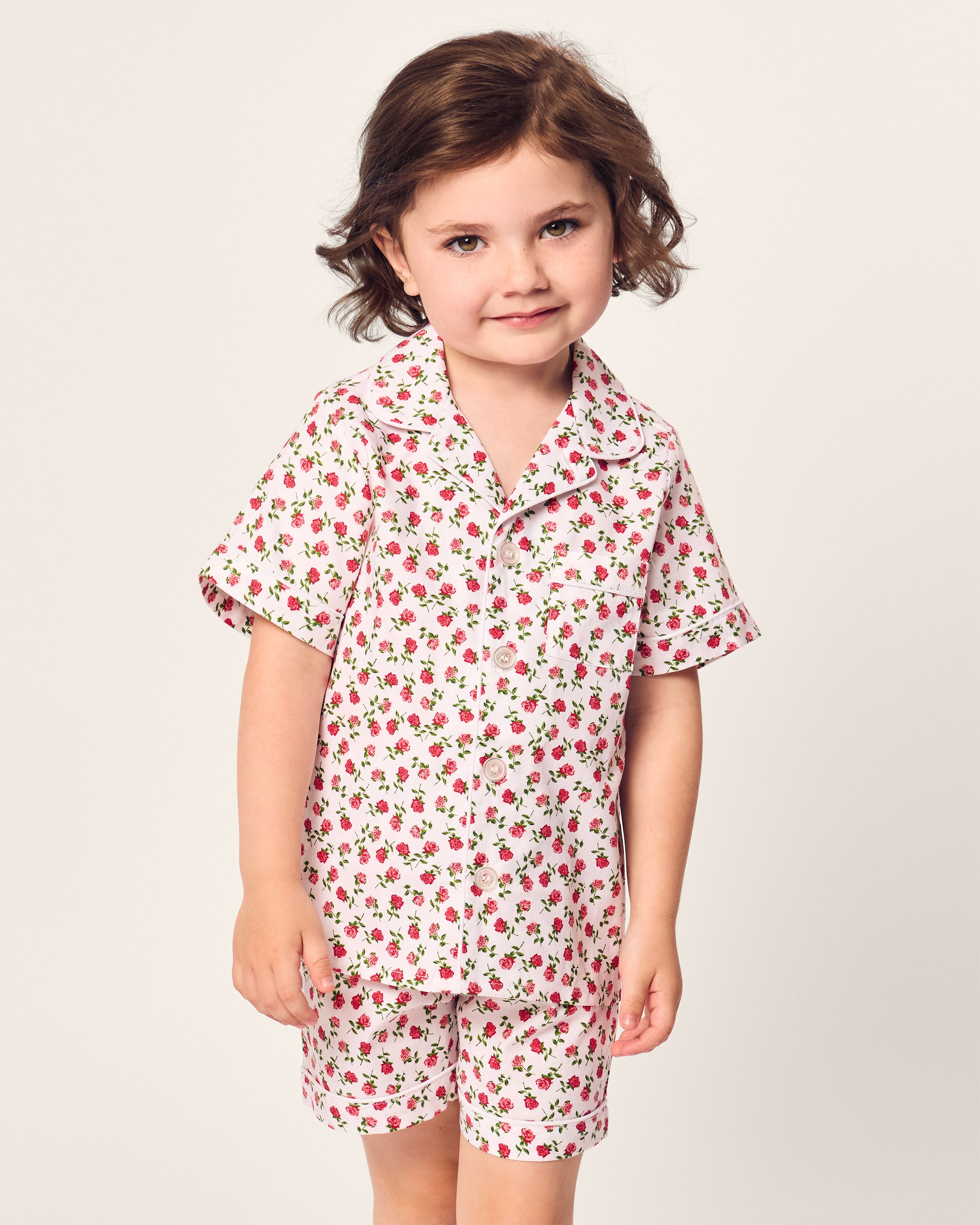 A child in Petite Plumes Kids Twill Short Set, featuring a fine cotton design with a red strawberry pattern, stands against a plain background, smiling slightly.