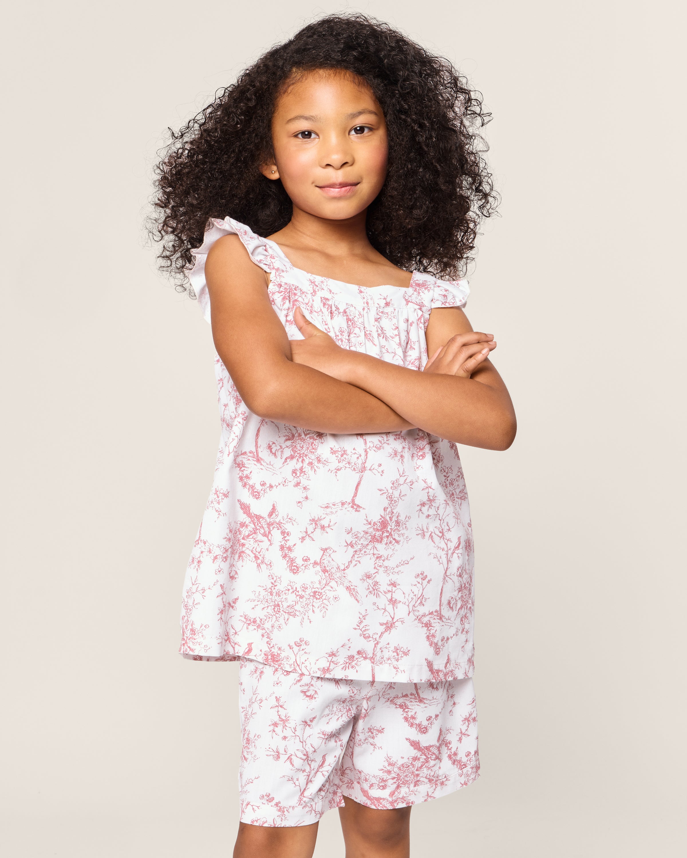 A young girl with curly hair stands with her arms crossed, dressed in Petite Plumes Girls Twill Clara Short Set in Pink Timeless Toile. The plain, light background showcases her delightful ensemble.