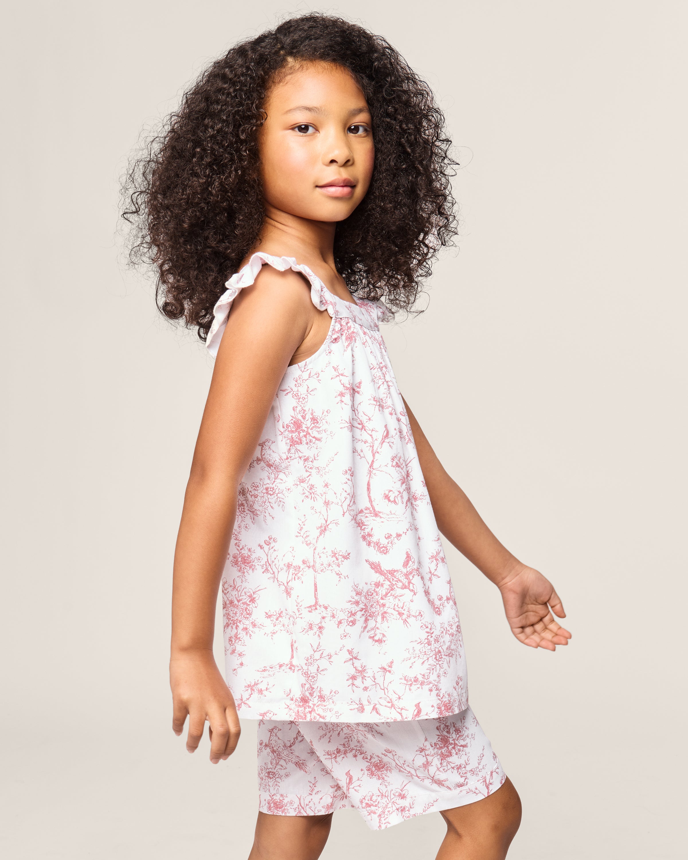 A young girl with curly hair wears the Girls Twill Clara Short Set in Pink Timeless Toile by Petite Plume, featuring a sleeveless white top with matching shorts in a pink floral pattern. She stands against a plain background, smiling slightly towards the camera.