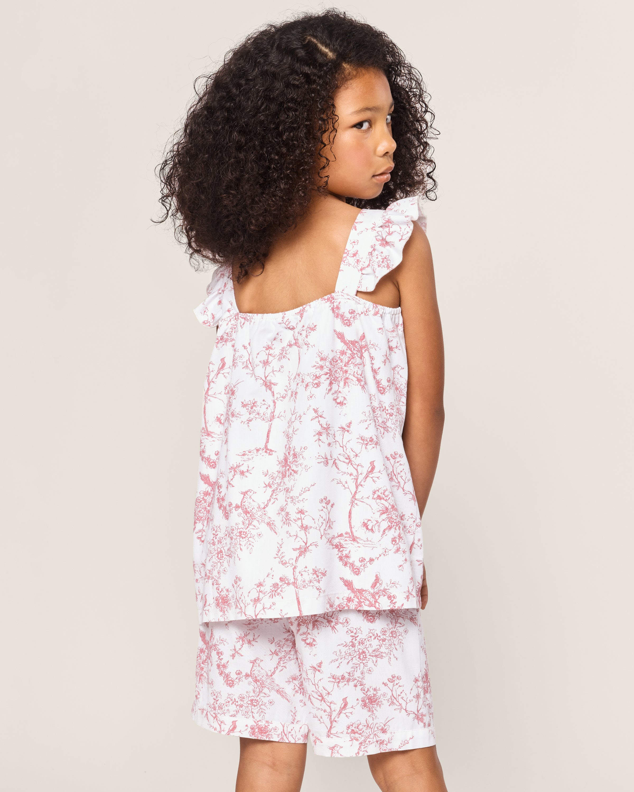 A curly-haired girl gazes over her shoulder, wearing a Petite Plume Girls Twill Clara Short Set in Pink Timeless Toile—a sleeveless dress with matching shorts—against a simple light background.