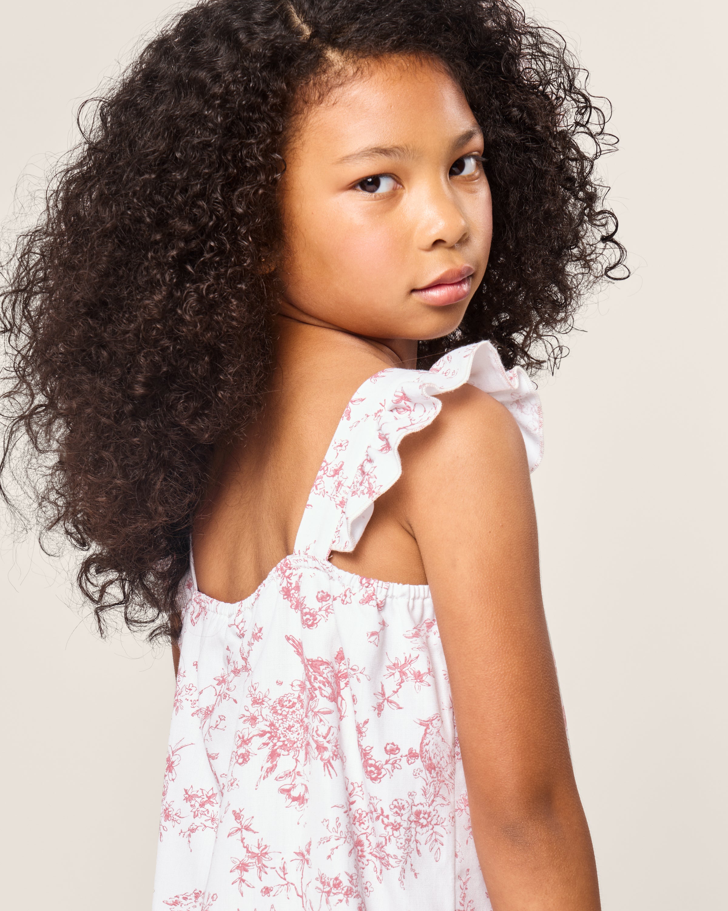 A young girl with curly hair looks over her shoulder, wearing Petite Plumes Girls Twill Clara Short Set in Pink Timeless Toile. The set, crafted from soft cotton sleepwear material, combines comfort and charm against the plain, light-colored background.