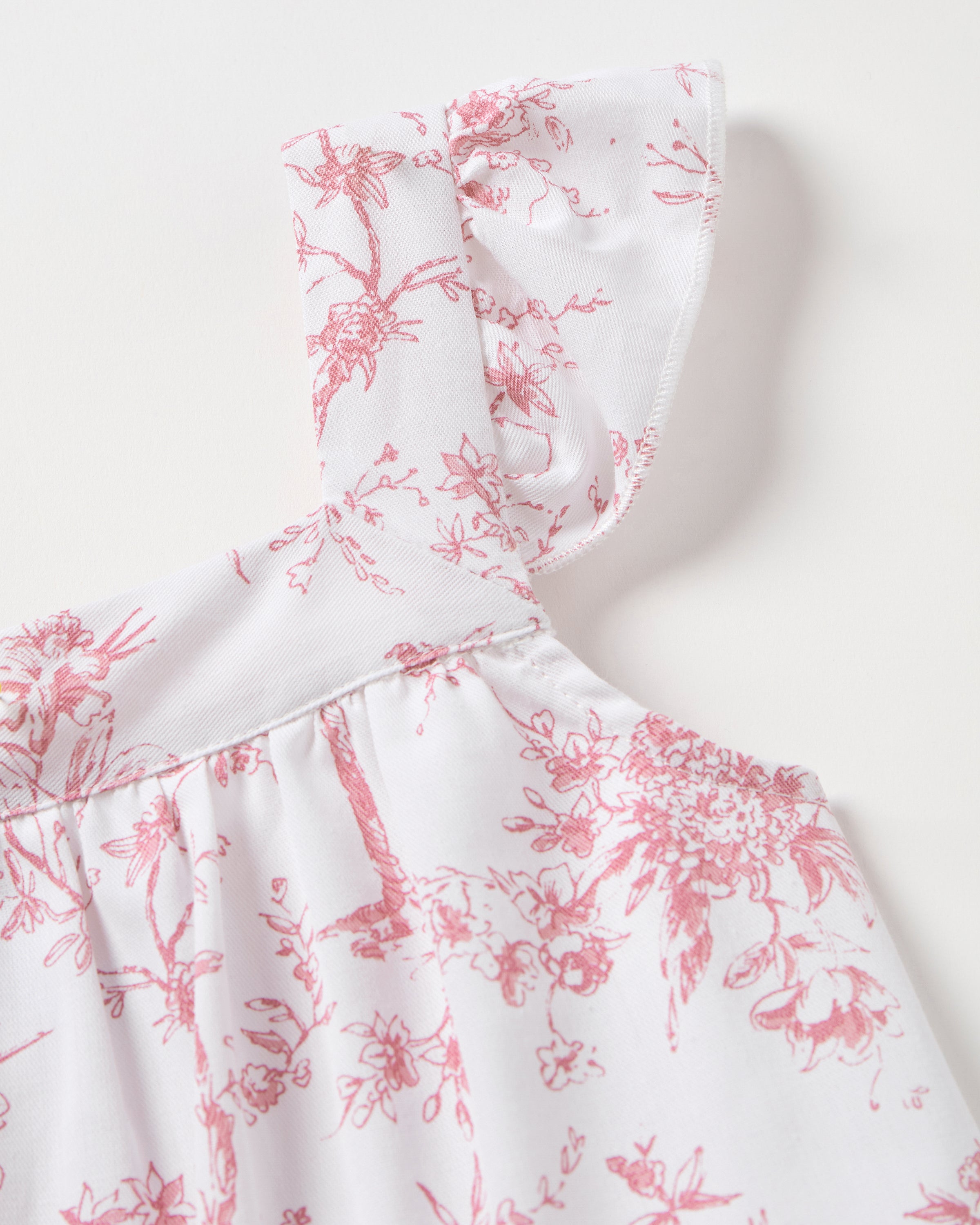 The Girls Twill Clara Short Set in Pink Timeless Toile by Petite Plume features delicate pink floral patterns on white fabric, a ruffled shoulder strap, and a gathered bodice, suggesting a feminine feel. The intricate design is evenly spread across the luxurious sleepwear piece.