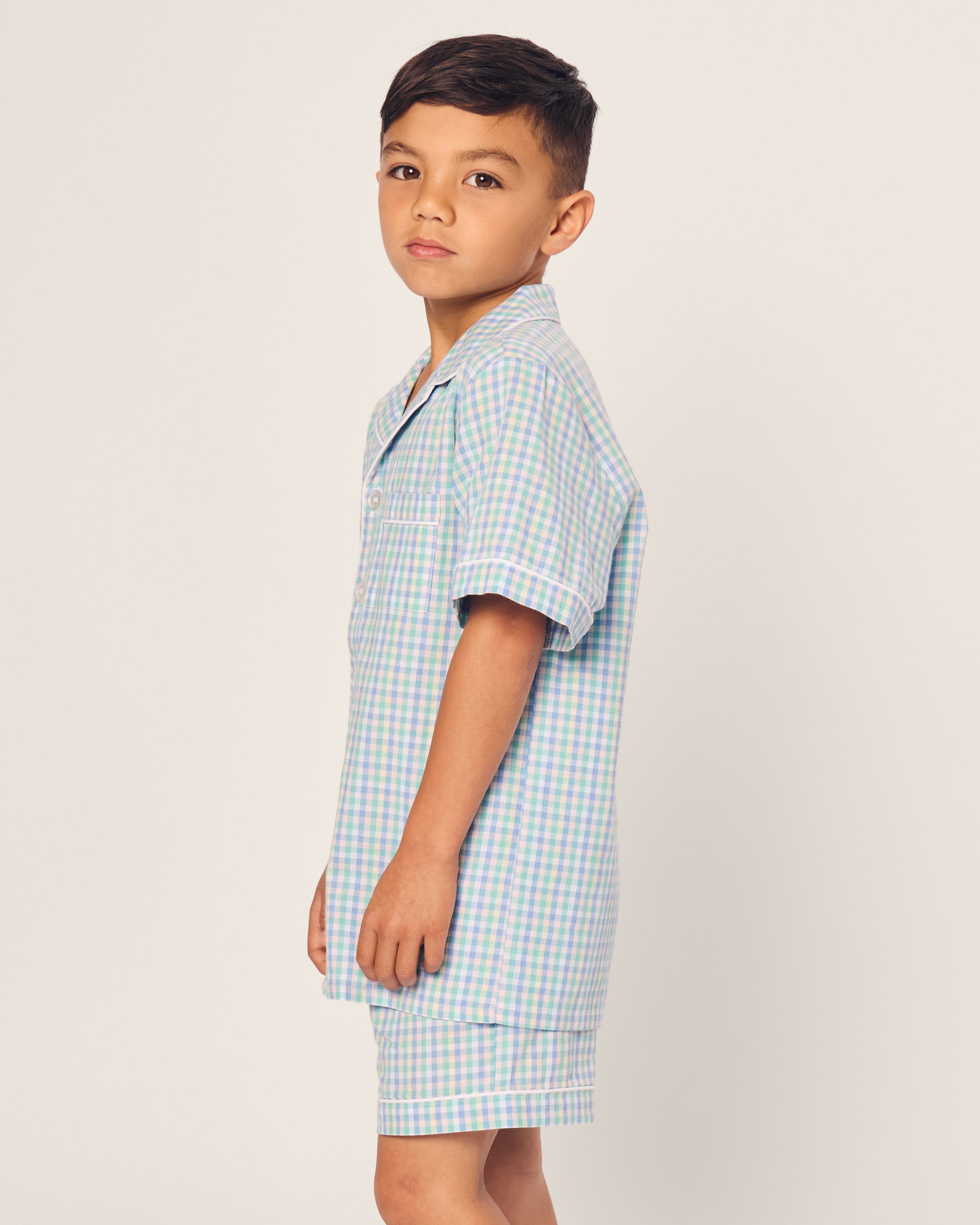 A young boy wears Petite Plumes Kids Twill Short Set in Spring Gingham, featuring a light blue and green plaid pattern. He stands sideways with a neutral expression against a plain light background.