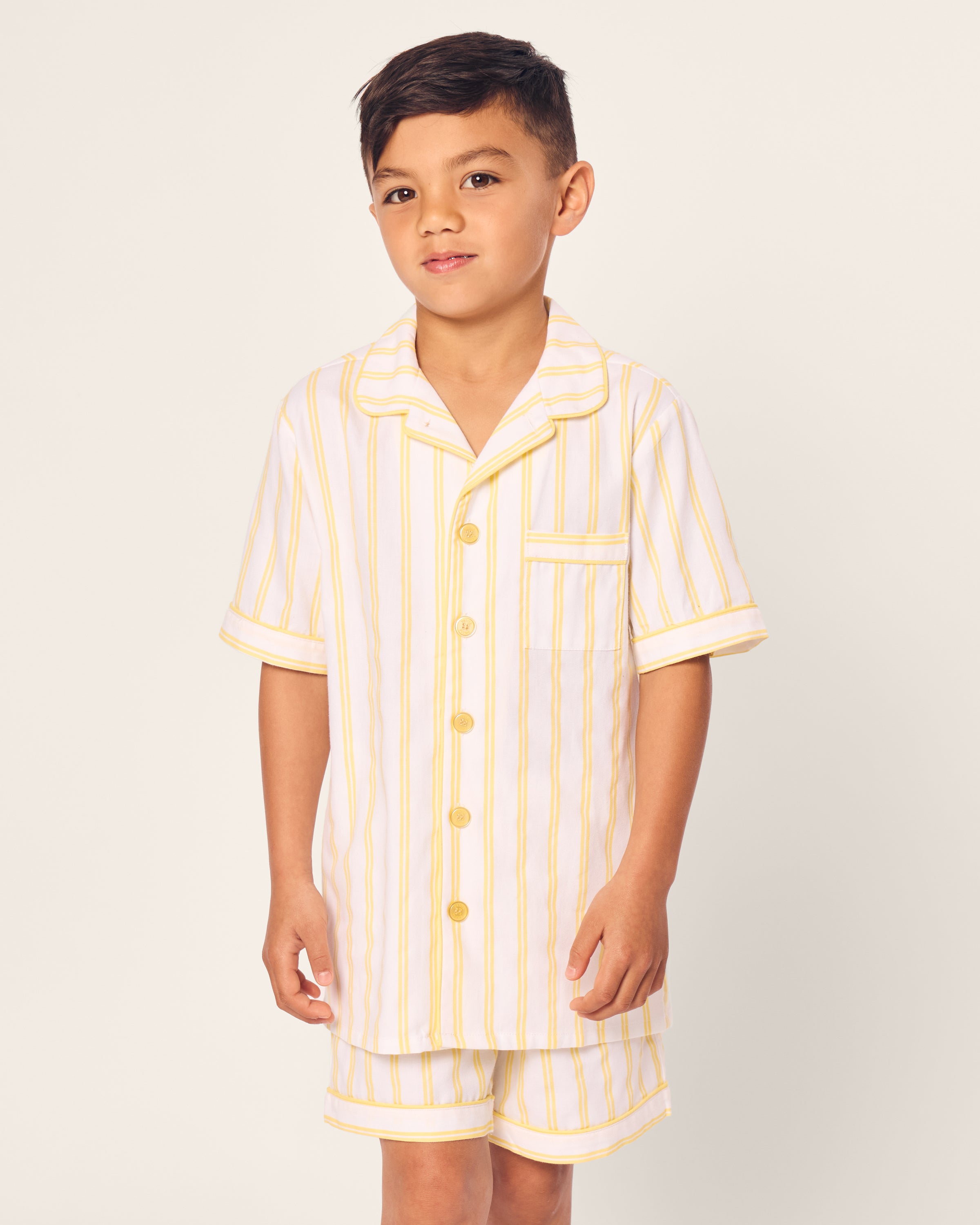 A young boy models the Kids Twill Short Sleeve Short Set in Sunny Stripe by Petite Plume, featuring flame-retardant cotton pajamas with white and yellow vertical stripes, a front pocket, and buttons, set against a simple light-colored background.