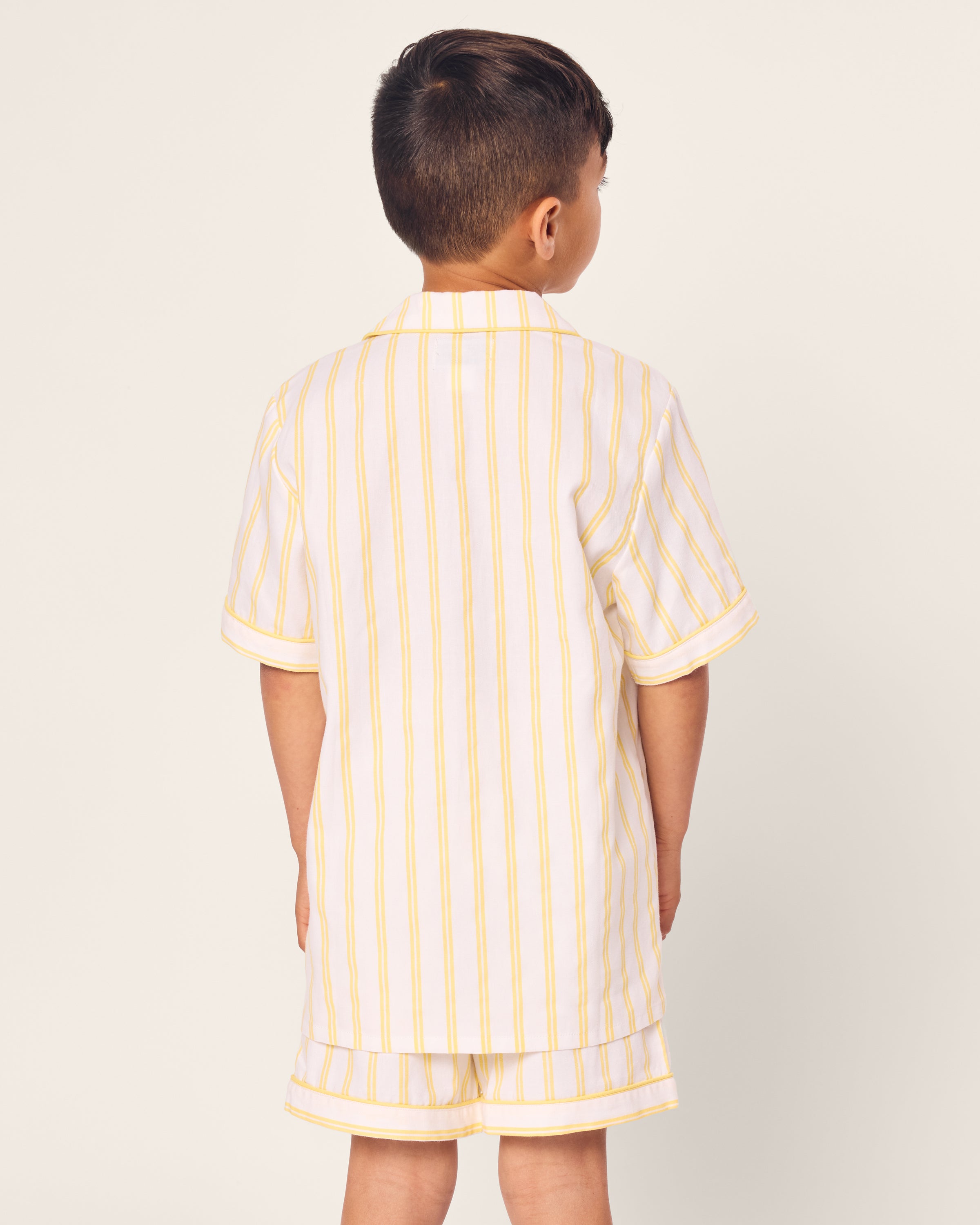 A young boy in Petite Plumes Kids Twill Short Sleeve Short Set in Sunny Stripe stands with arms relaxed by his sides against an off-white background.