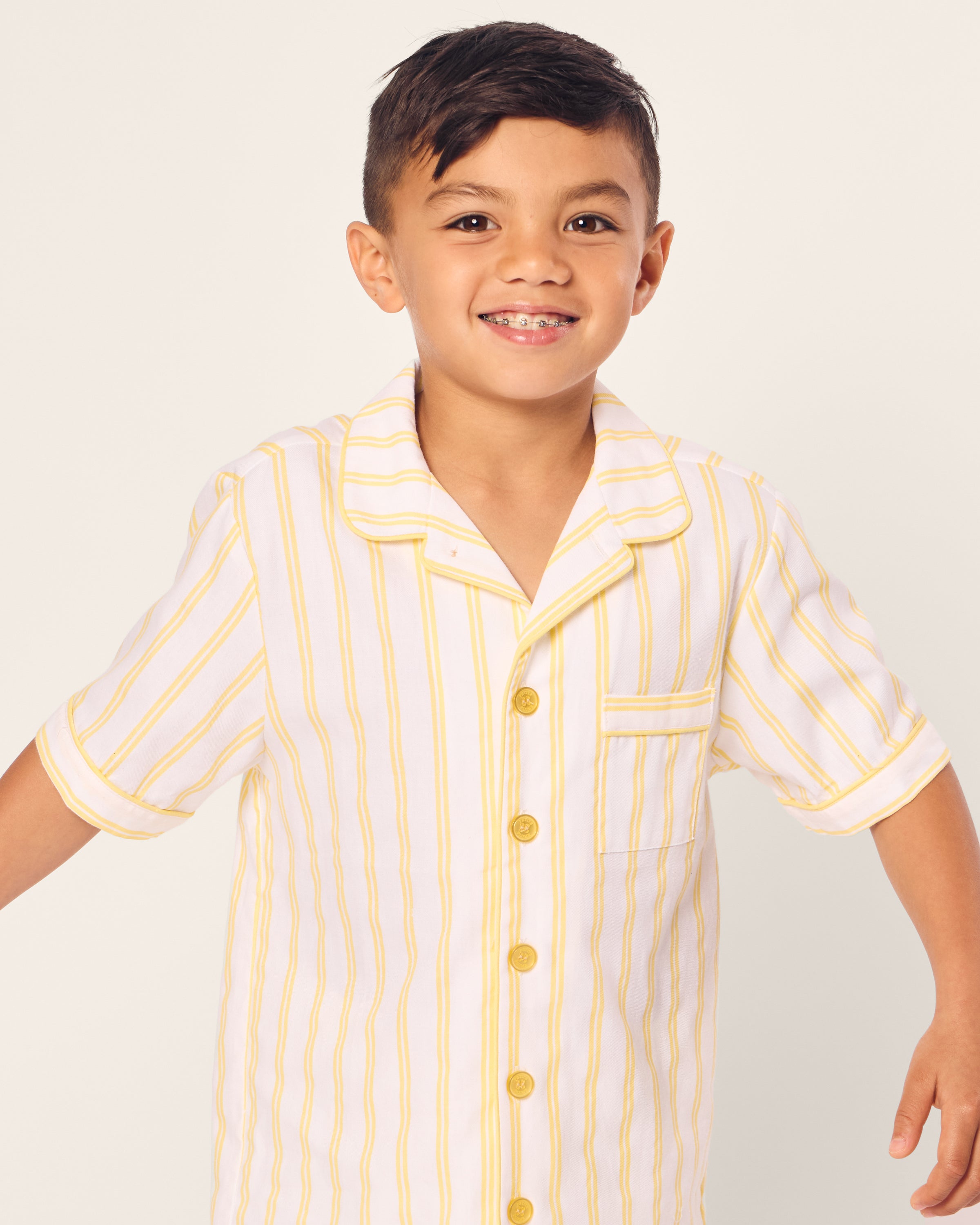 A young boy with a short haircut smiles, wearing the Petite Plume Kids Twill Short Sleeve Short Set in Sunny Stripe, featuring yellow vertical stripes and a pocket. The background is plain and light-colored.