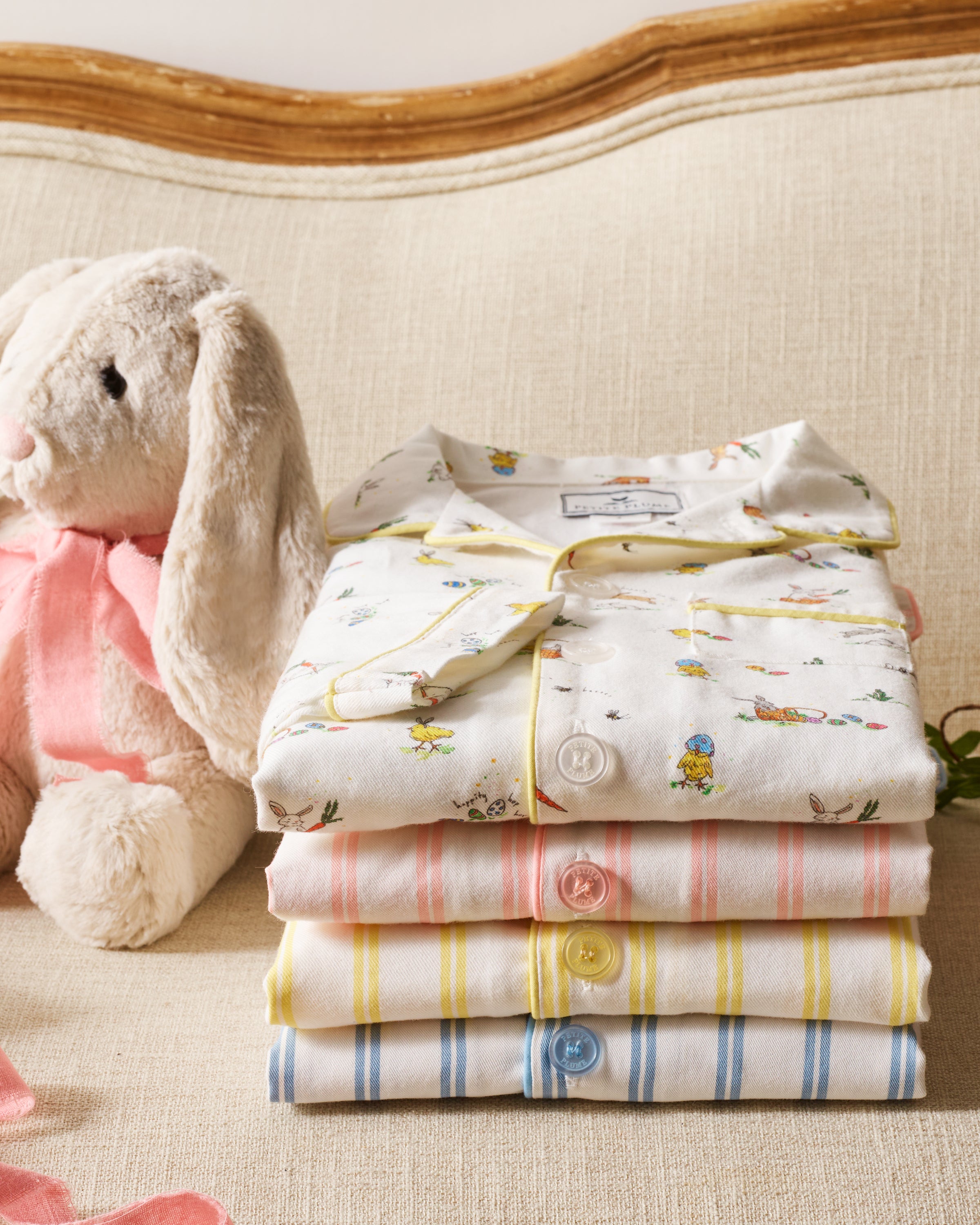 A stack of Petite Plumes Kids Twill Short Sleeve Short Sets in Sunny Stripe, made from soft, flame-retardant cotton for safety and comfort, sits neatly on a beige sofa. To the left, a plush white bunny with a pink bow rests against light wooden-accented sofa.