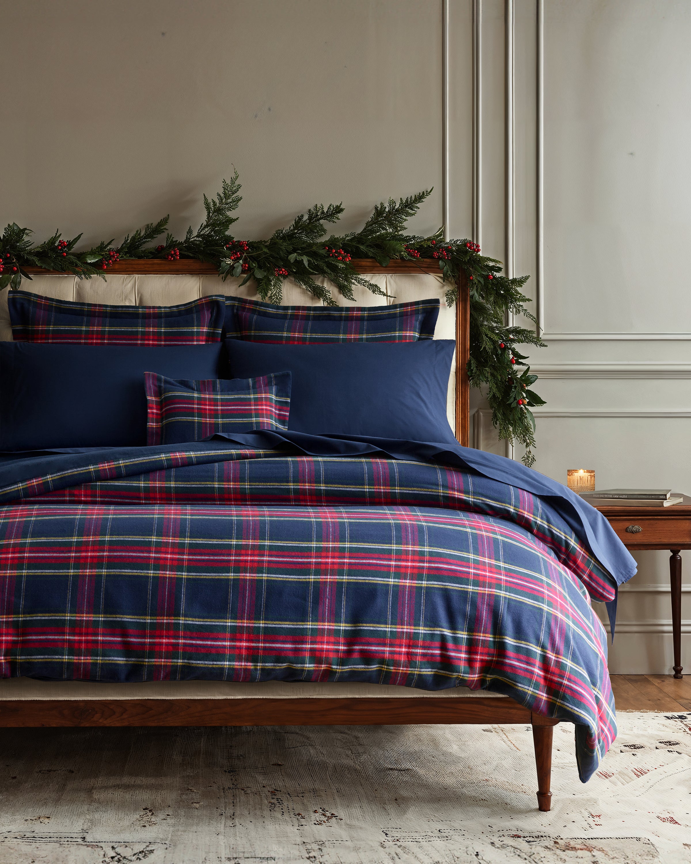 A bedroom features a neatly made bed with the Petite Plume Flannel Duvet Set in Windsor Tartan. The headboard is adorned with green garland and red berries, enhancing the winter bedding theme. A wooden side table with a lit candle completes the cozy setting against light-colored walls.