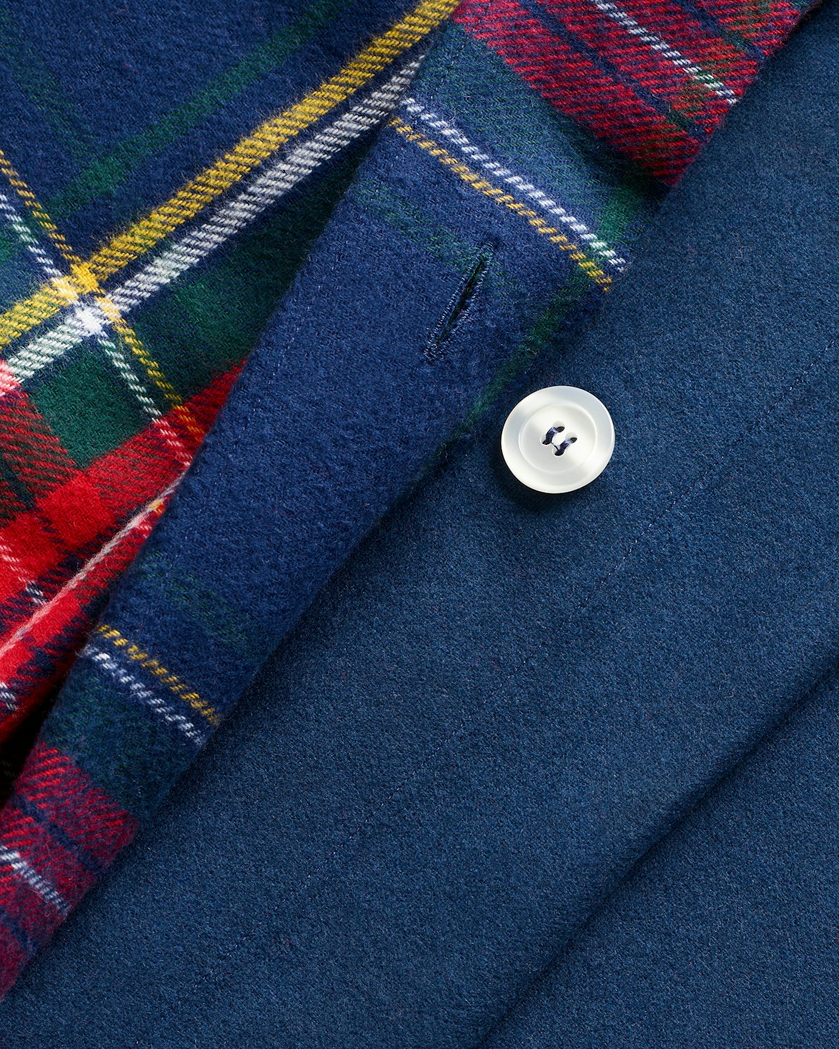 Close-up of a blue wool blazer with a single white button, revealing a fabric underneath reminiscent of the Flannel Duvet Set in Windsor Tartan by Petite Plume. The red, green, and blue plaid pattern adds cozy winter bedding vibes to the composition.