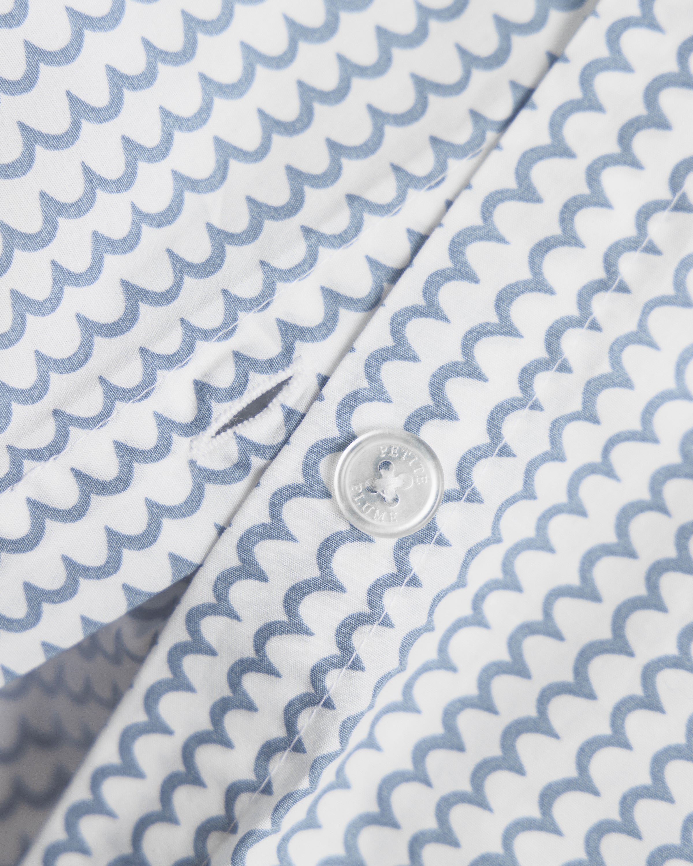 A close-up of the Petite Plume Luxe Premium Percale Duvet Set in La Mer shows a white shirt with a repeating blue scallop design, 100% cotton. It has clear round buttons and neat buttonhole stitching, with fabric as soft and smooth as luxury sheets.