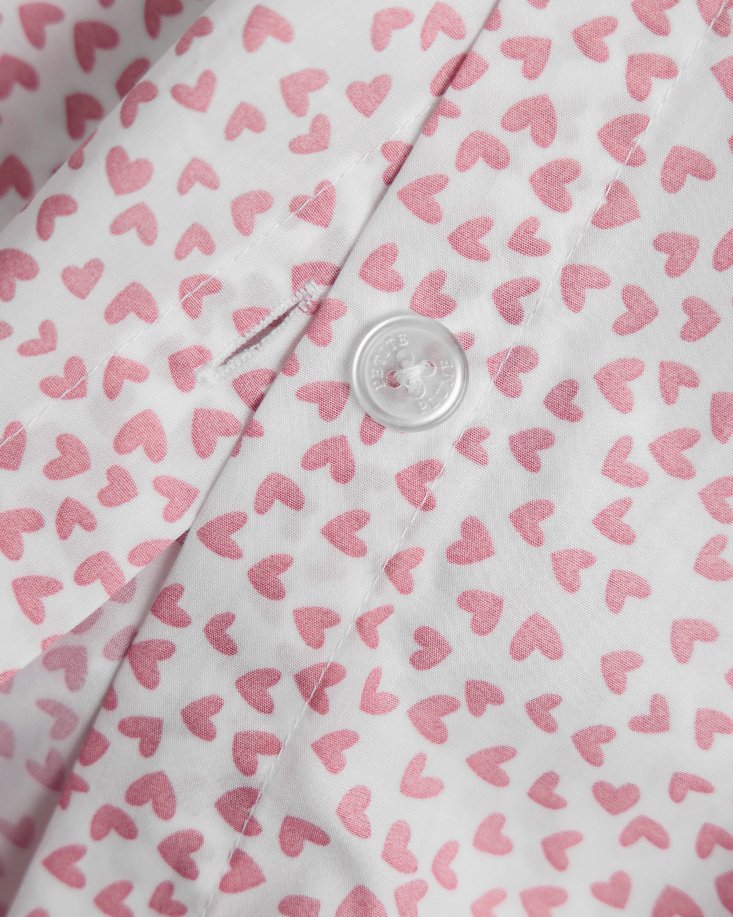 Close-up of the Luxe Premium Percale Duvet Set in Sweethearts by Petite Plume, featuring 100% cotton fabric adorned with small pink hearts and a white button at the center. Visible stitching and a buttonhole near the button suggest its part of an elegant bedding ensemble.