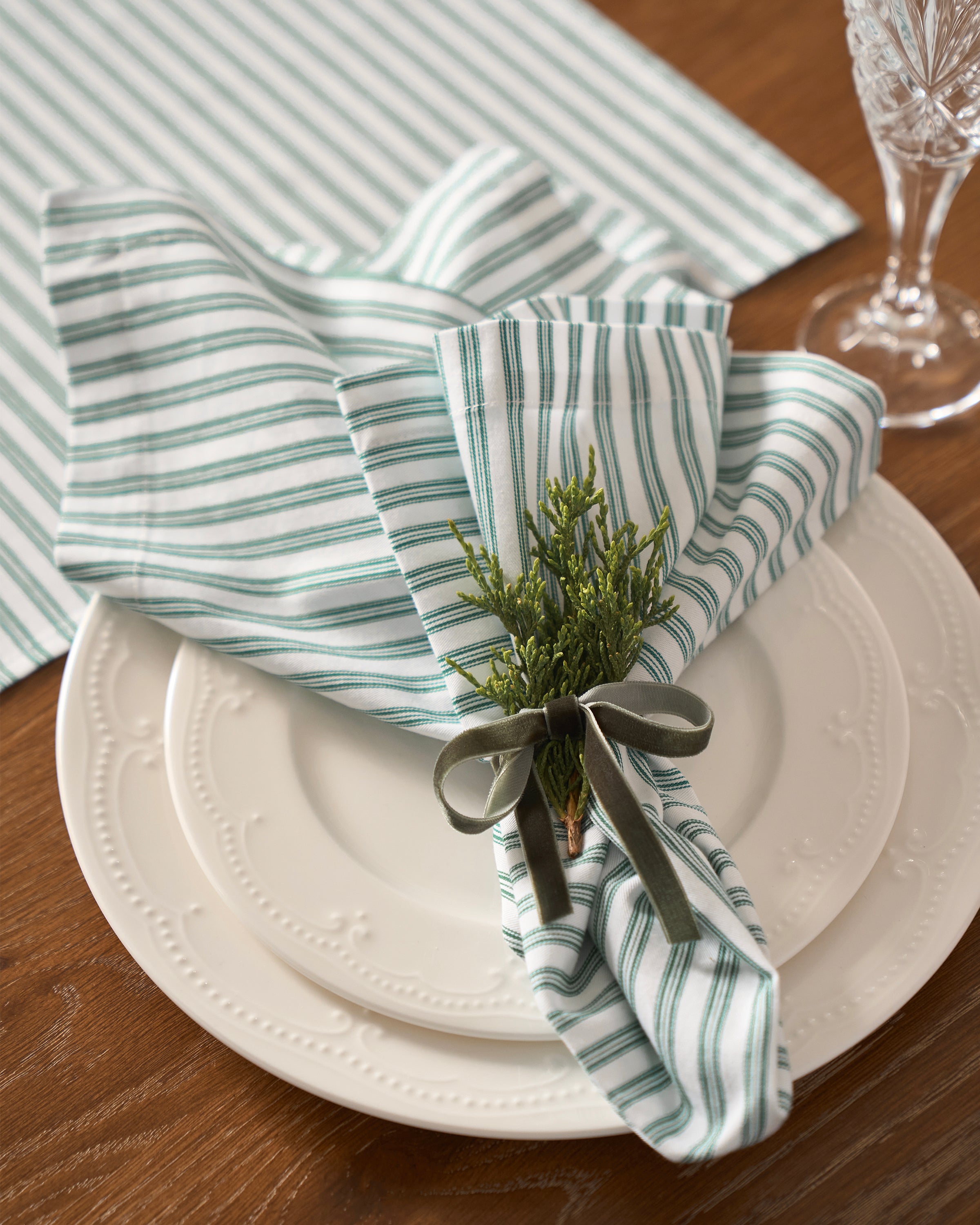 A 100% cotton, intricately designed plate rests on a wooden surface. Petite Plumes Signature Twill Napkins in Emerald Ticking are elegantly folded with a sprig of greenery and secured with a green ribbon, enhancing the festive tablescape. A crystal glass sparkles nearby.