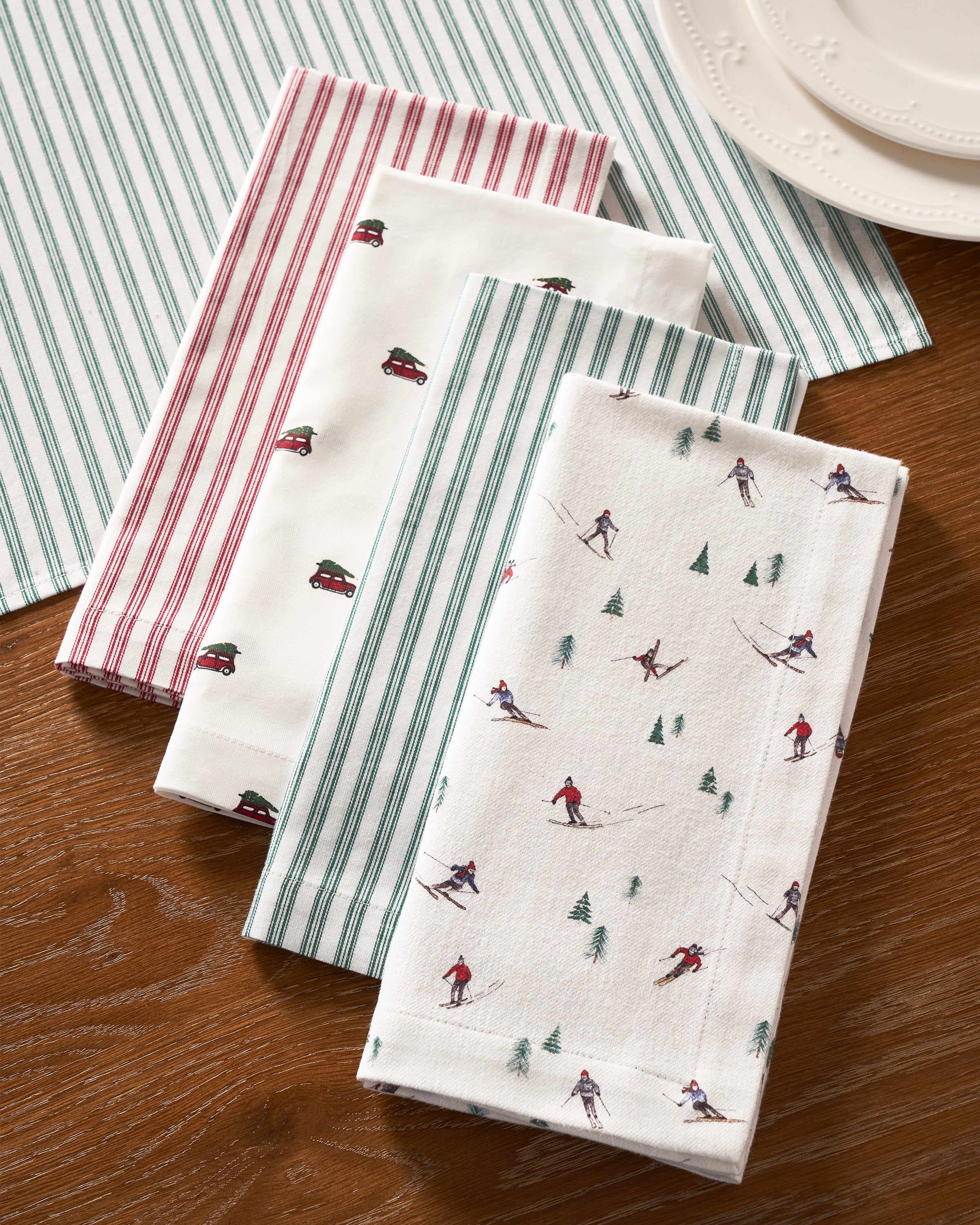 Four Signature Twill Table Napkins by Petite Plume are arranged on a wooden table, featuring designs of red stripes, green stripes, small red cars with green trees, and skiers with pine trees. Stacked white plates are partially visible in the top right corner, enriching the festive tablescape.