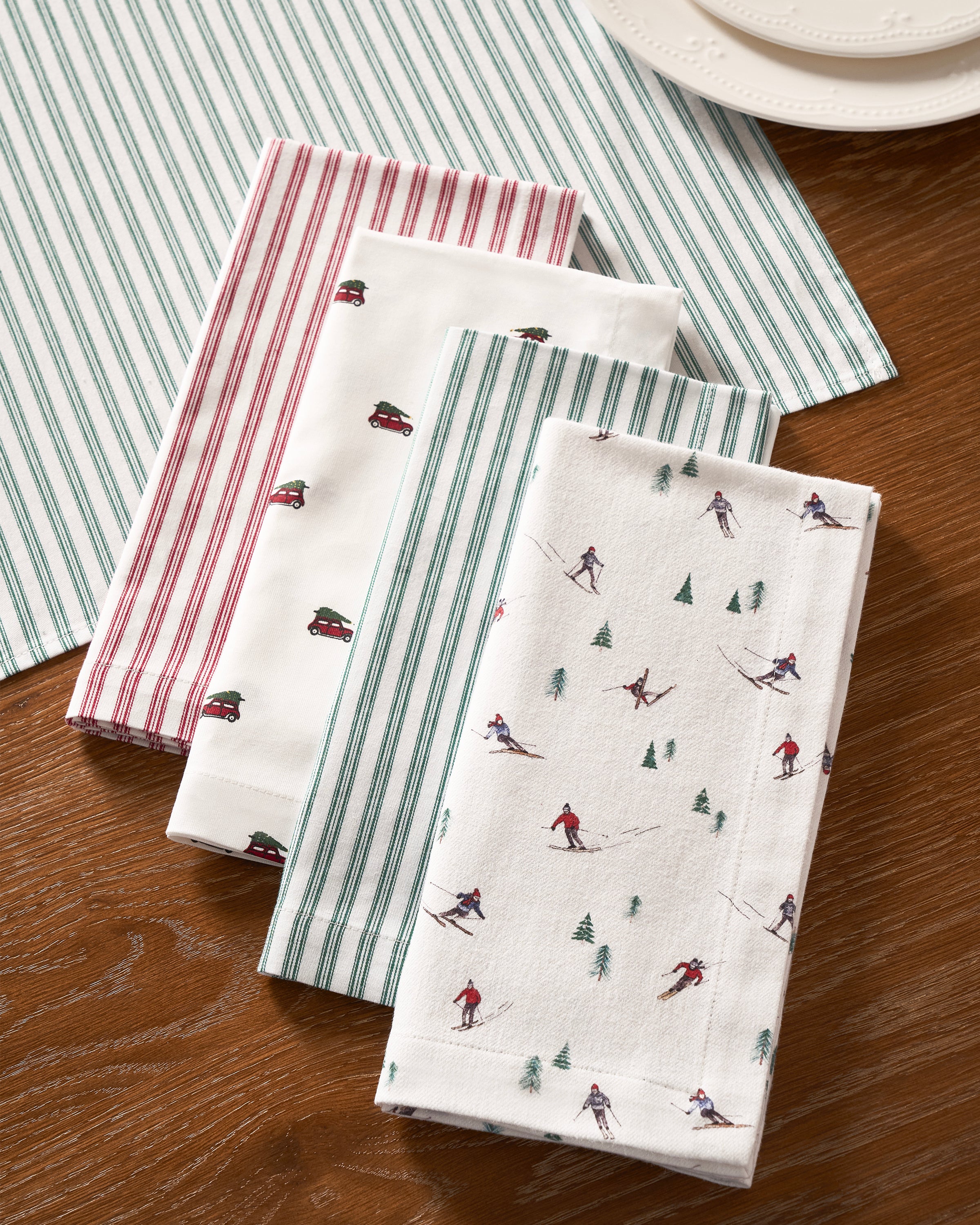 Four Signature Twill Napkins in patterns (red stripes, vintage cars, green stripes, skiing figures with trees) by Petite Plume sit on a wooden table. Beneath them is a matching striped placemat, and a white ornate plate peeks out from the background.