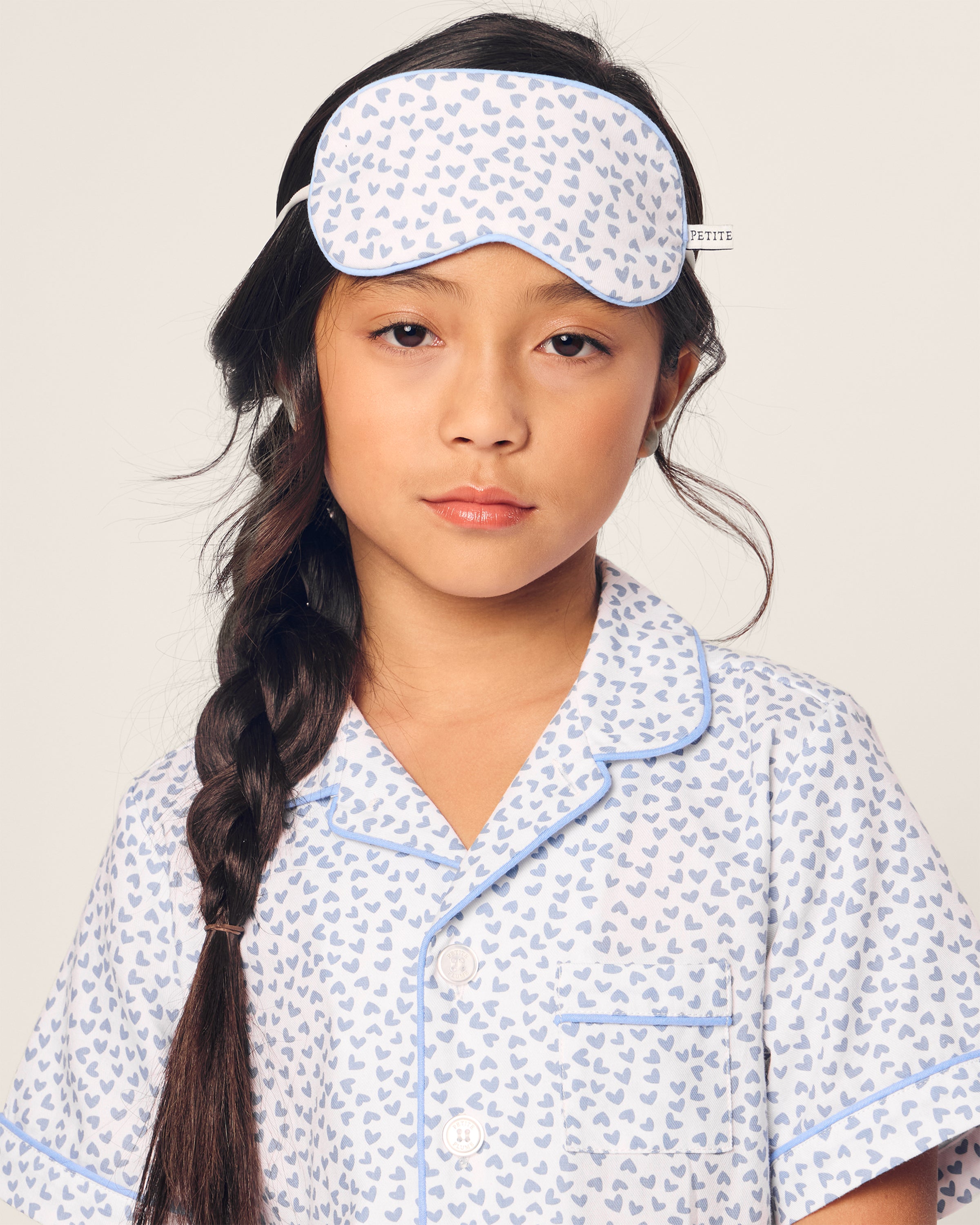 A child with long hair in a braid wears Petite Plumes Kids Twill Sleep Mask in the Bluehearts design, complementing the charming pajama set with stylish blue piping and buttons, creating an enchanting bedtime look against the plain white background.